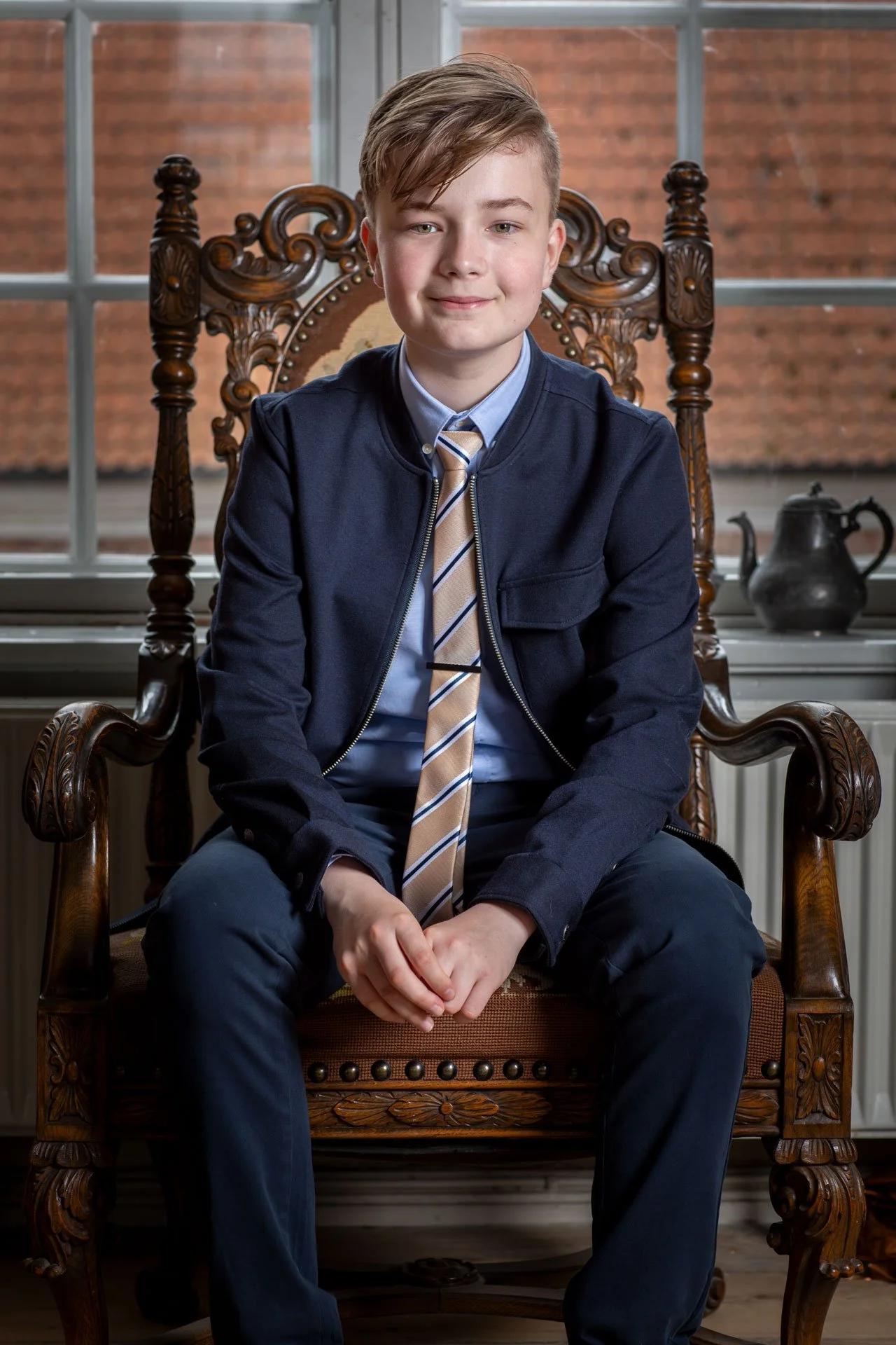 En ung dreng sidder i en stor, gammel træstol foran et vindue med murstensvæg baggrund. Han er klædt i konfirmationstøj med blazer, skjorte, slips og bukser.