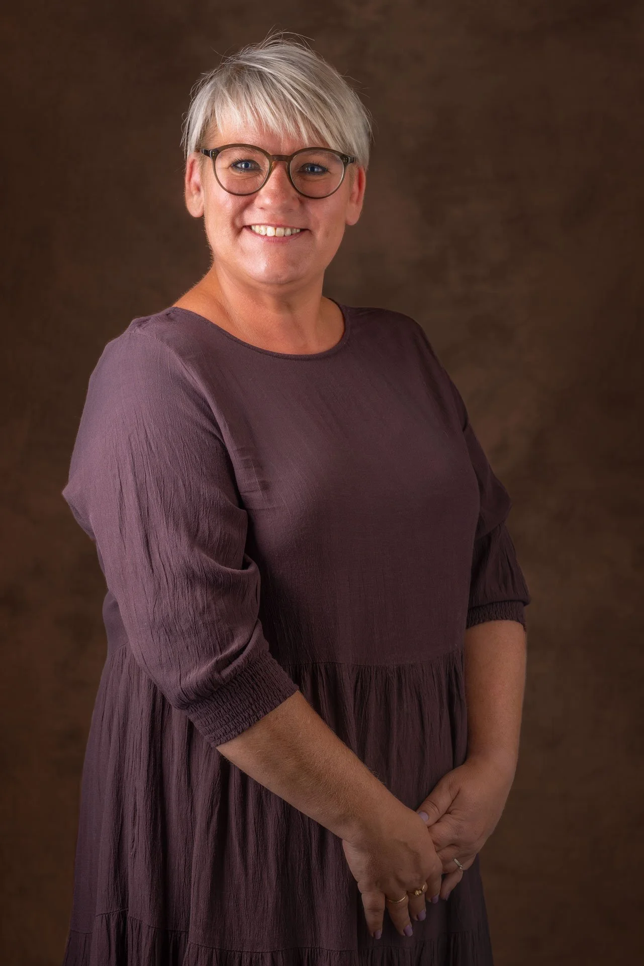 Portræt af en kvinde med blond, kort hår, briller, smil, iført en lilla kjole, stående mod en brun baggrund.