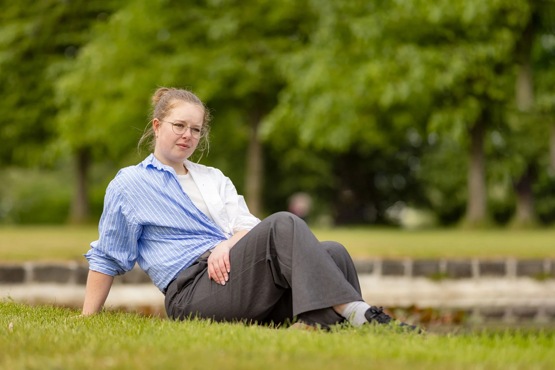 En ung kvinde sidder på græsset i en park med grønne træer bag sig.