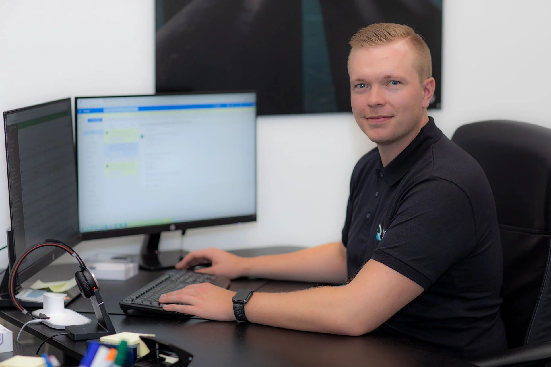 En mand sidder ved et skrivebord med to computer screens, et tastatur og en mus, og smiler mod kameraet.