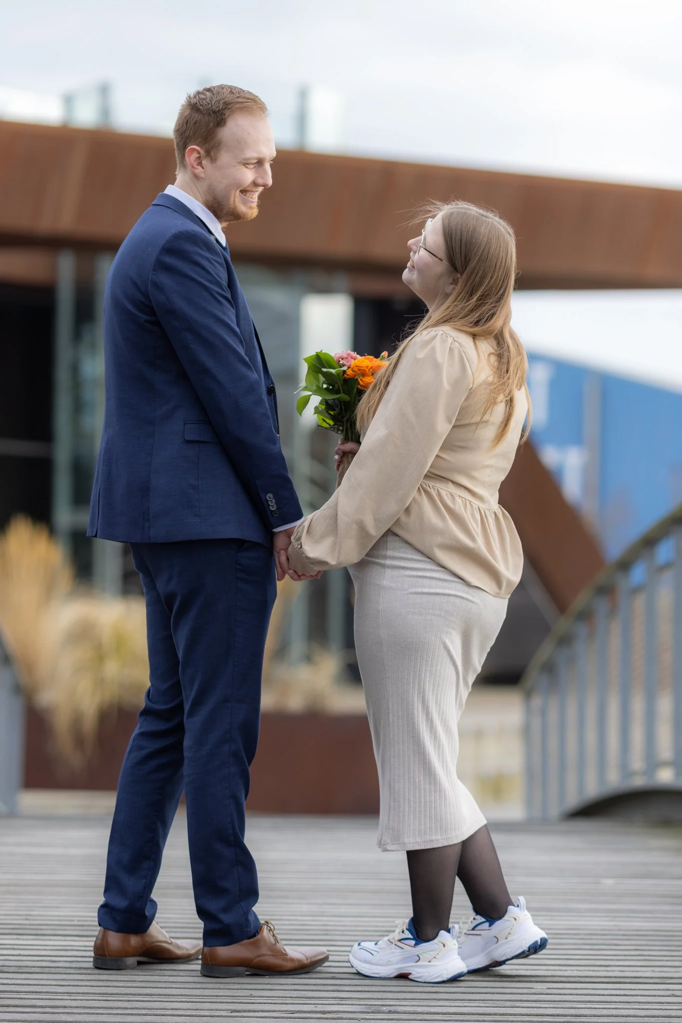 En mand og kvinde står ansigt til ansigt på en træplankebrygge, holder i hænder og ser på hinanden, kvinden holder en buket blomster.