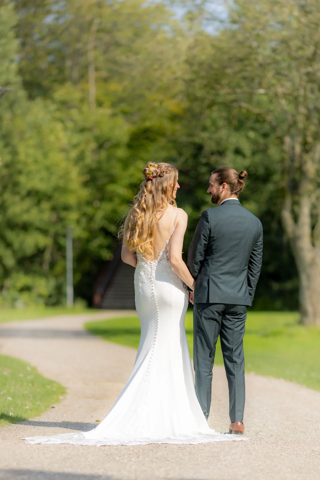 Et brudepar står på en sti i en grøn park, holder hinanden i hånden og kigger på hinanden.