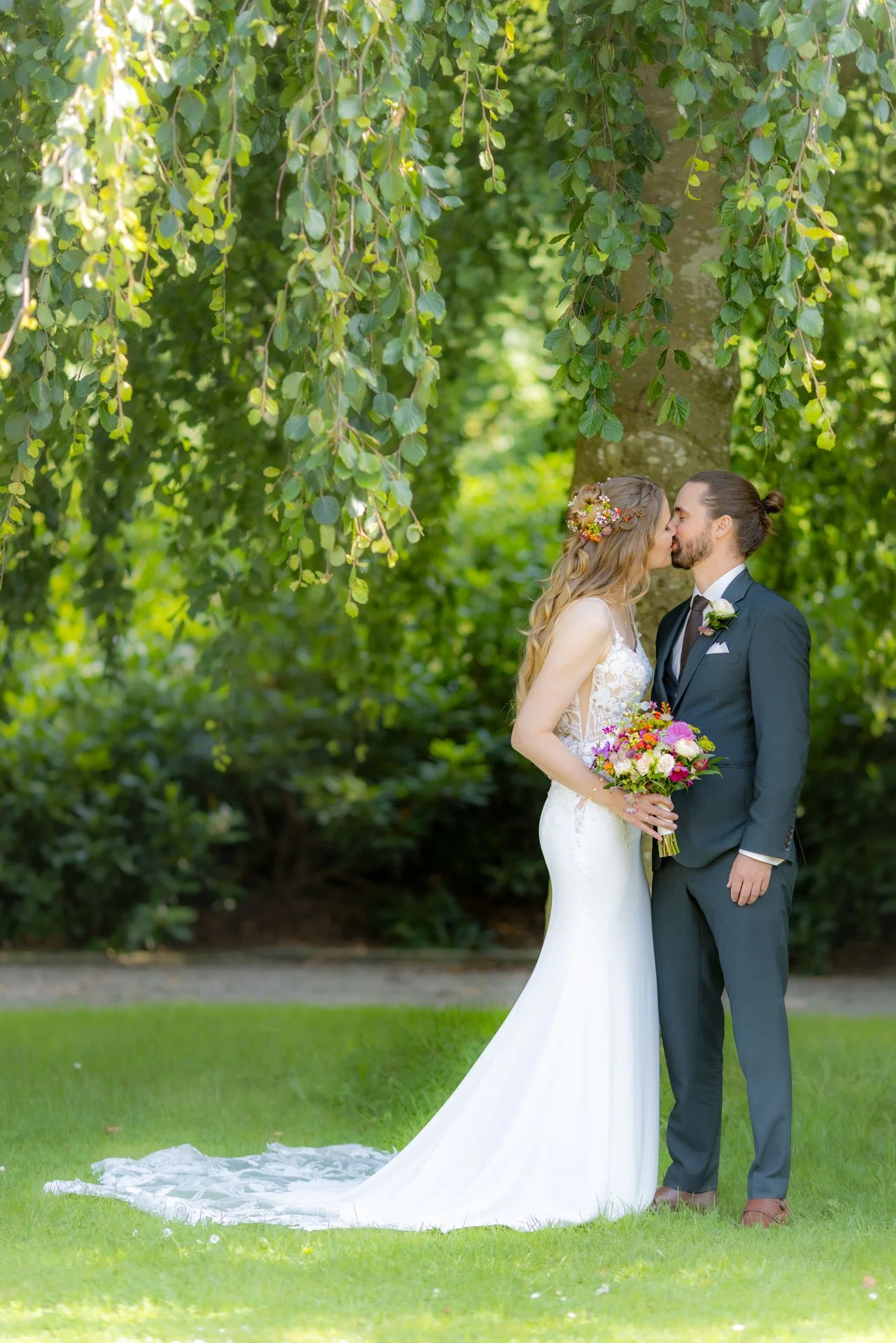 Par, der kysser i en park med grønne træer. Kvinden bærer en hvide brudekjole med blomsterranke i håret og holder en farverig blomsterbuket; manden har tøj i mørk farve og et hvidt skjortetilbehør.