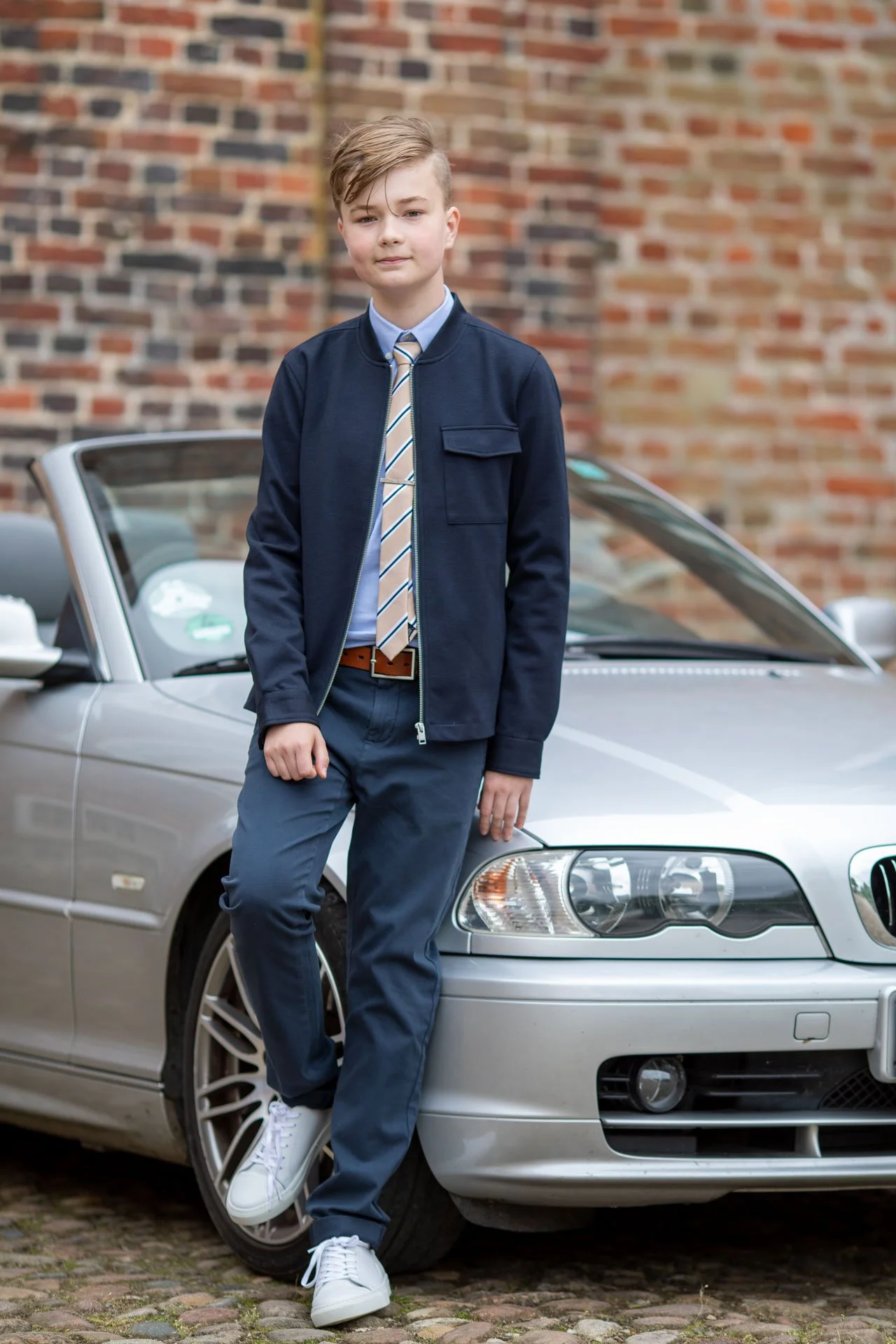 En ung dreng står foran en sølvfarvet sportsvogn, der står på brosten, foran en murstensvæg.