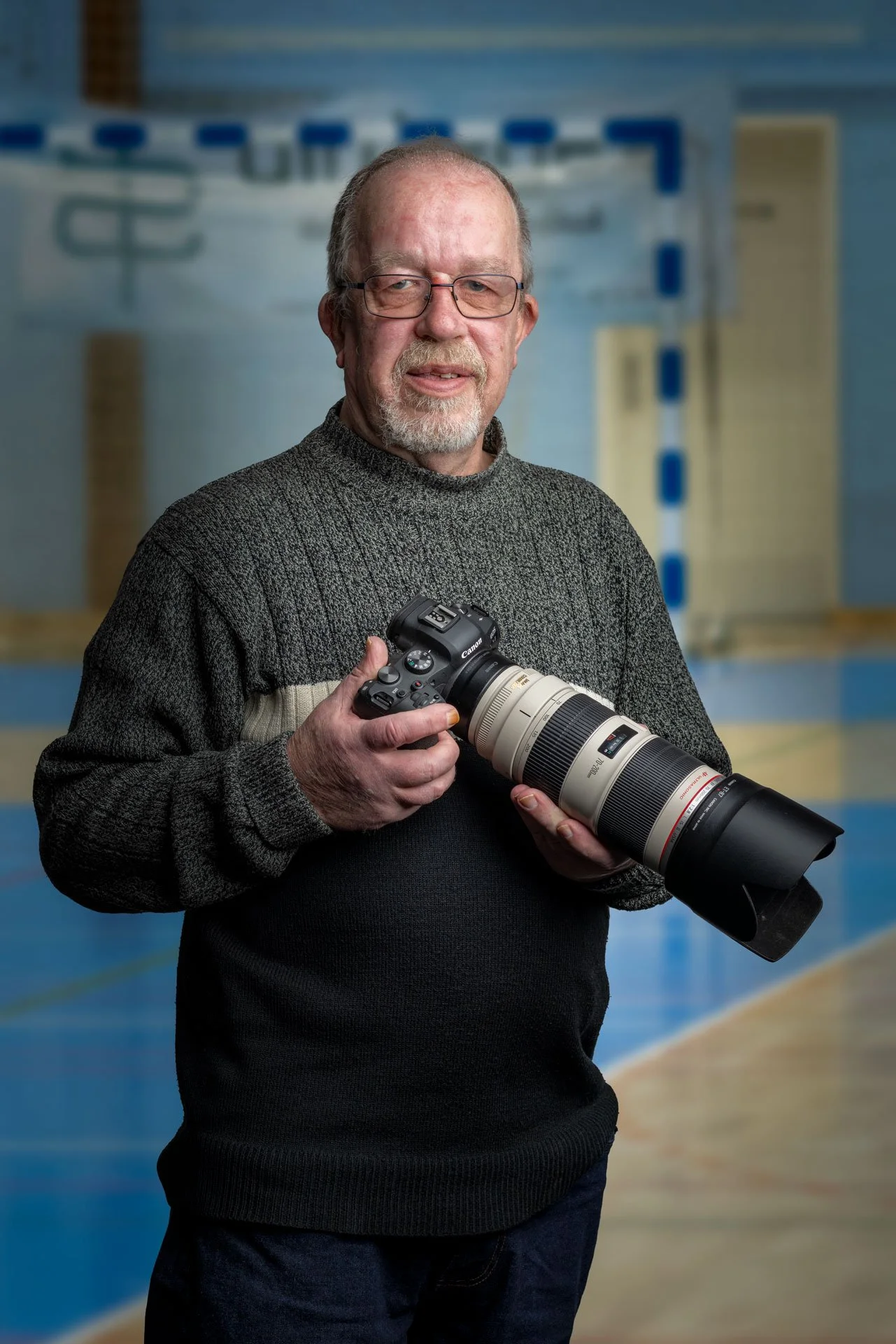 En ældre mand med brunt hår og skæg, iført en grå sweater, står i en gymnastiksal og holder et professionelt kamera med teleobjektiv.