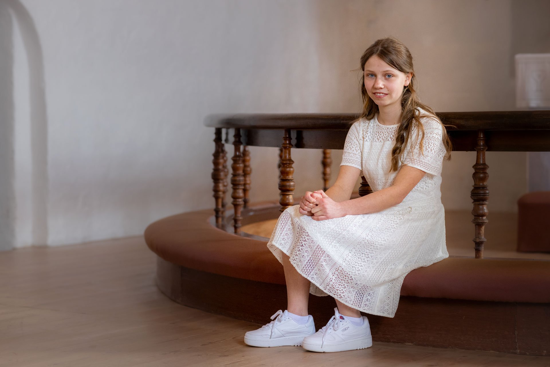 Ung pig sidder på en rund, træbænk indendørs, iført en hvid, konfirmaionskjole og hvide sneakers, med hænder foldet i skødet.