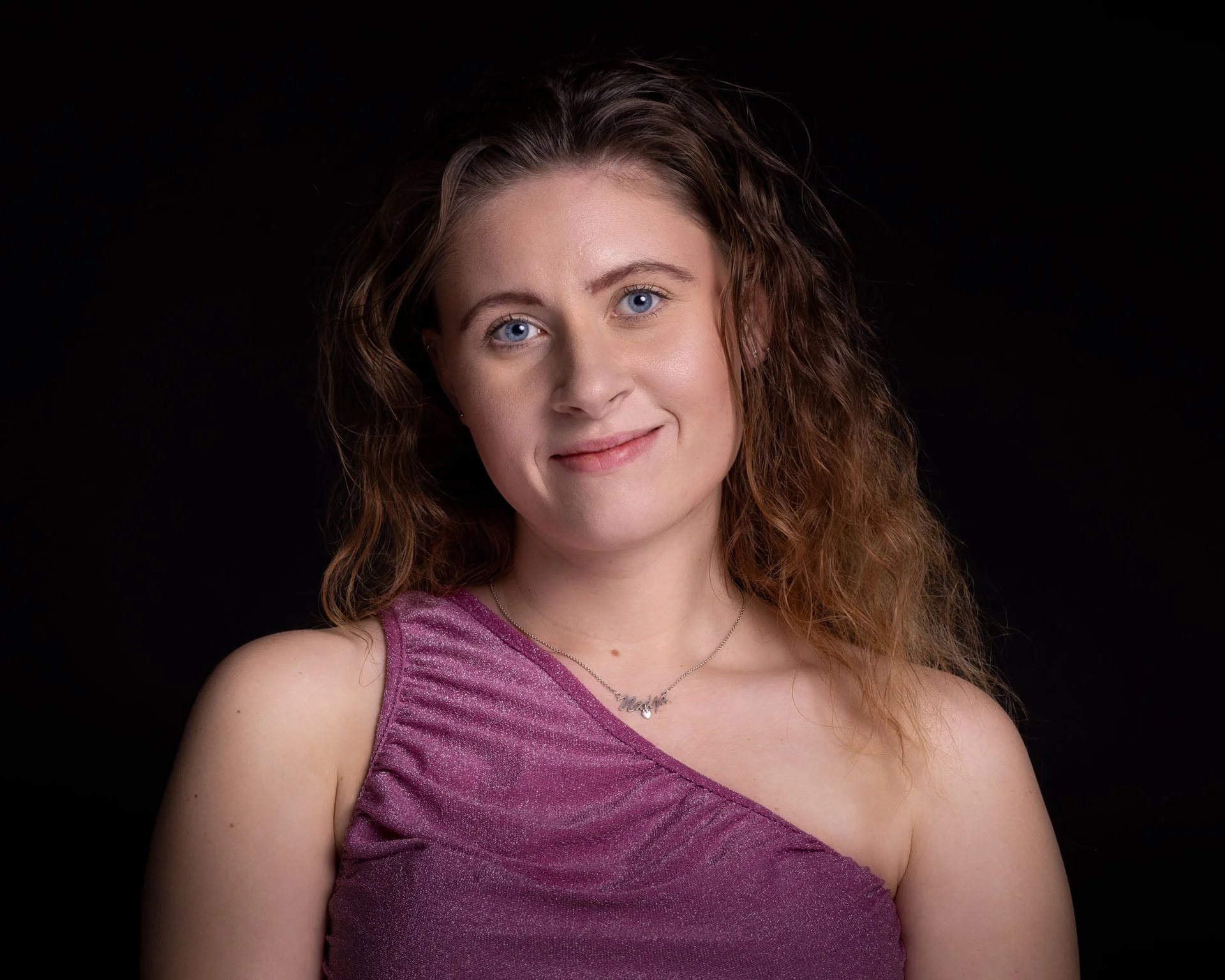 Portræt af en ung kvinde med brunt, krøllet hår iført en lilla, enkelt stof top, smilende mod kameraet mod en sort baggrund.