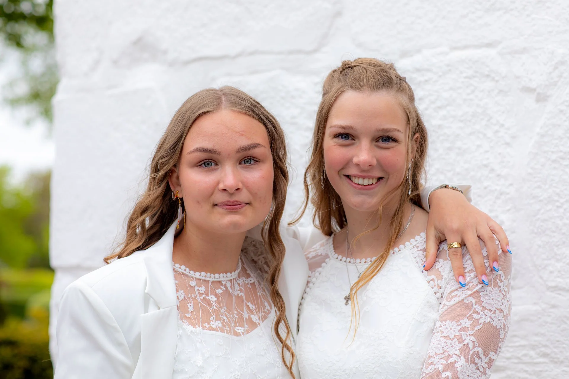 To unge kvinder iklædt hvide kjoler står tæt sammen foran en hvid væg, og smiler til kameraet.