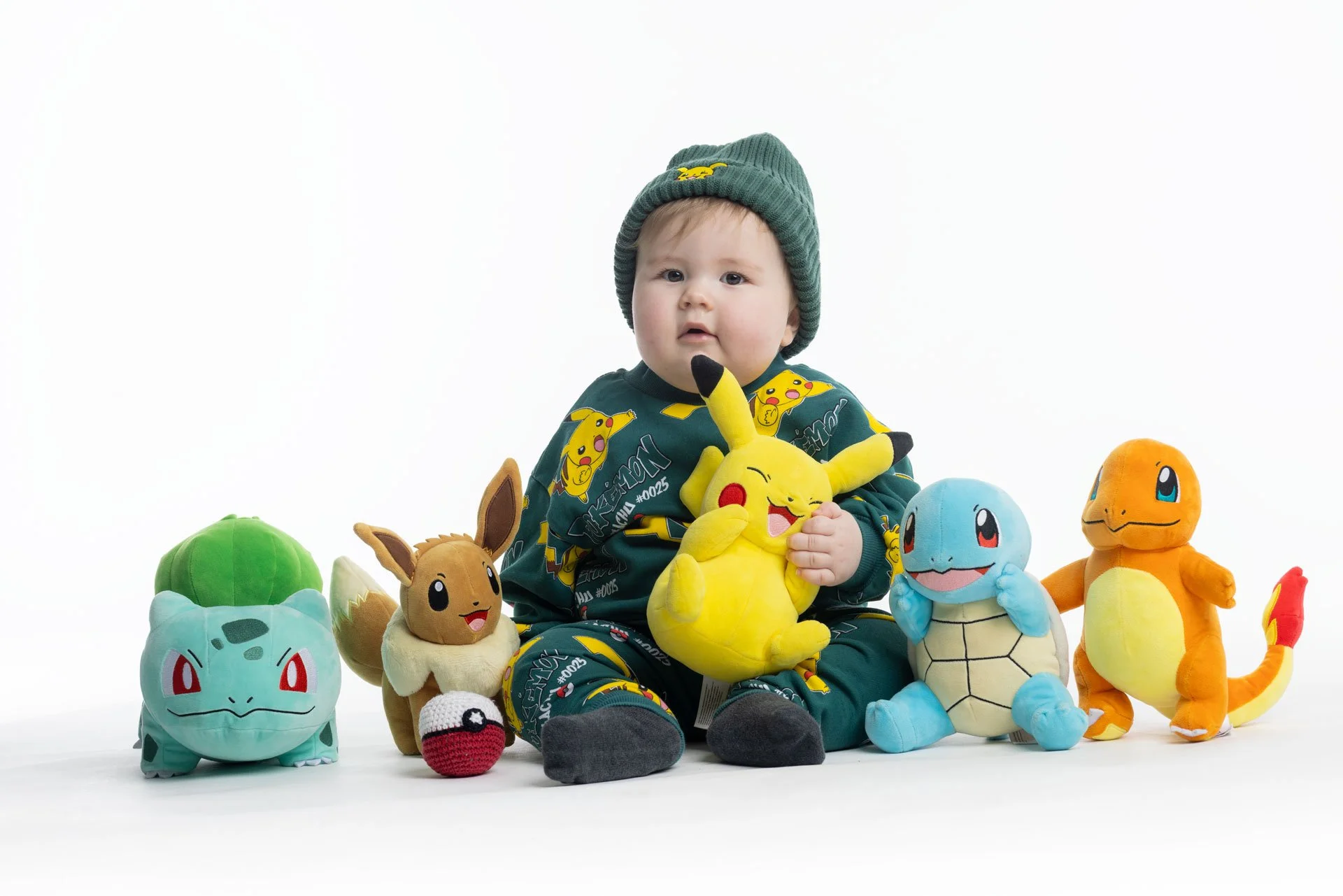 Børneunge sidder omgivet af Pokémon-dukker, herunder Pikachu, Bulbasaur, Eevee, Squirtle, Charmander, og et Pokémon-æg, på en hvid baggrund.