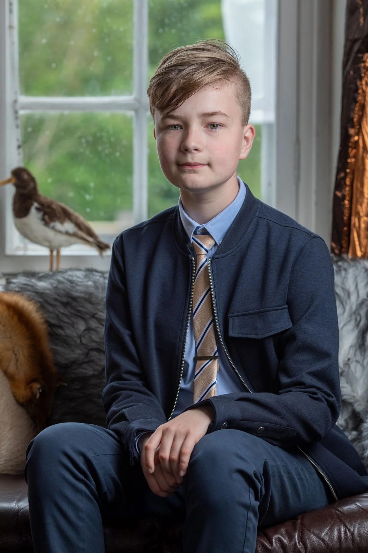 En ung dreng sidder på en sofa foran et vindue med grønne træer udenfor. Der er en fugl og en hund i baggrunden.