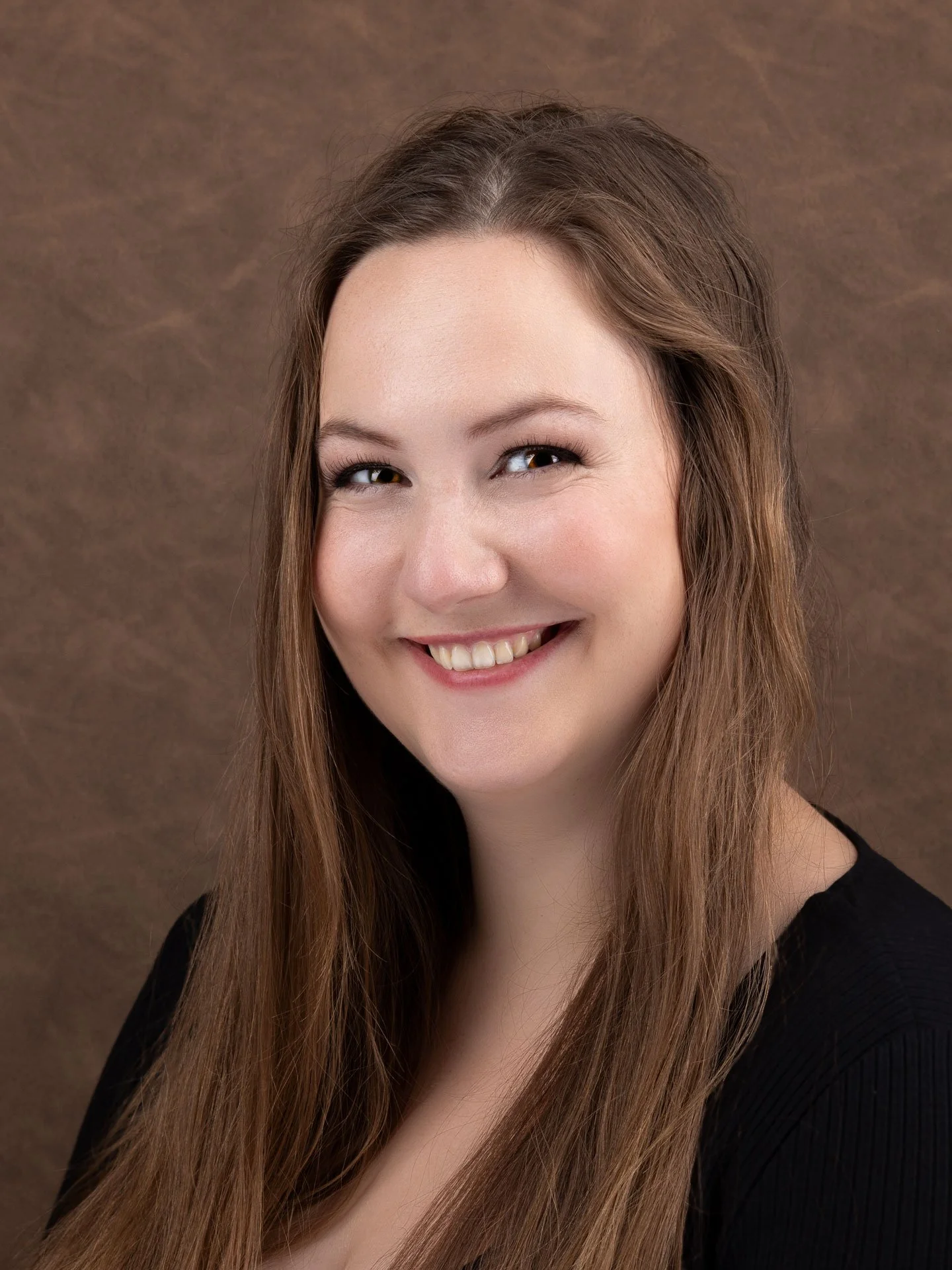 Et smilende portræt af en ung kvinde med langt, brunt hår og sort top mod en brun baggrund.
