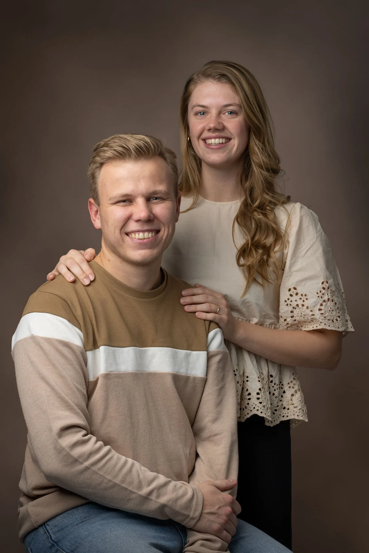 Et portræt af en ung mand og en ung kvinde, hvor kvinden står bag manden med hånden på hans skulder, begge smiler mod kameraet mod en neutral baggrund.