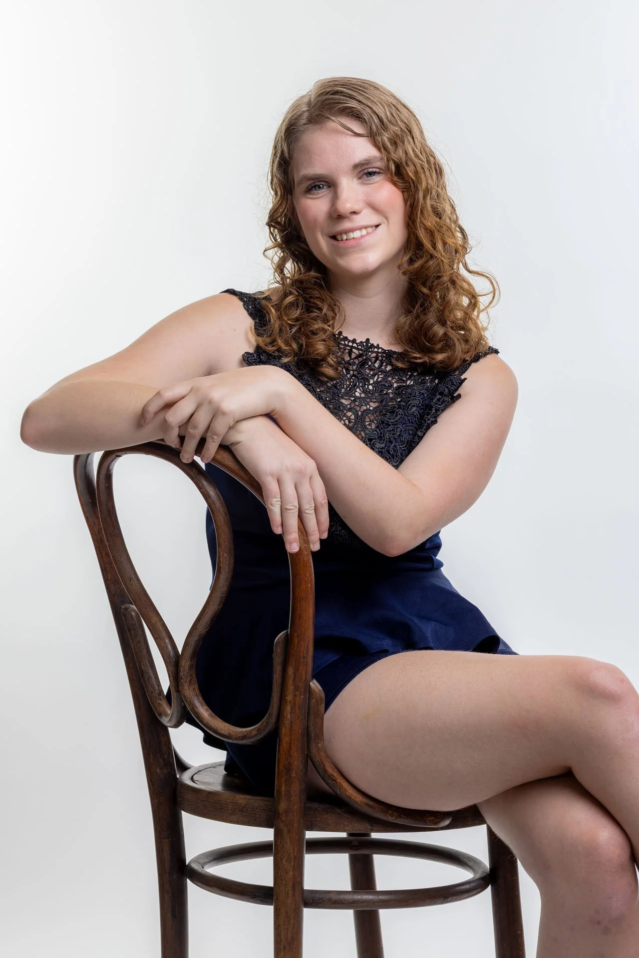 En kvinde med krøllet, mellemlangt hår smilende, iført sort blondertop og mørk blå nederdel, sidder på en træstol mod en hvid baggrund.