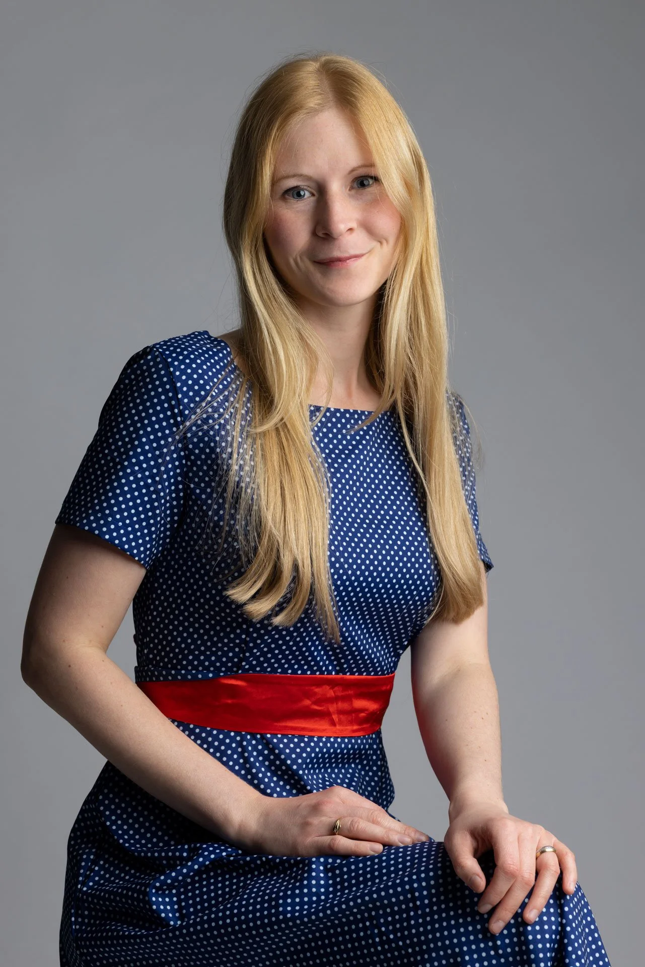 En kvinde med langt blondt hår, iført en blå kjole med hvide prikker og en rød talje, sidder mod en grå baggrund.