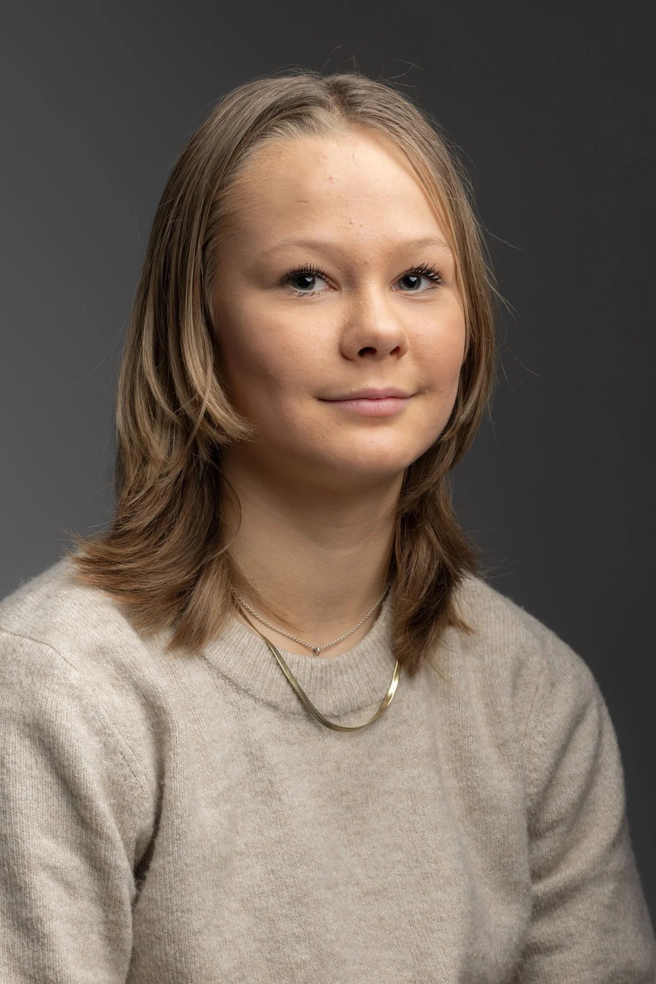 Portræt af en ung kvinde med skulderlangt brunt hår, iført en beige sweater og smykker, mod en mørk grå baggrund.