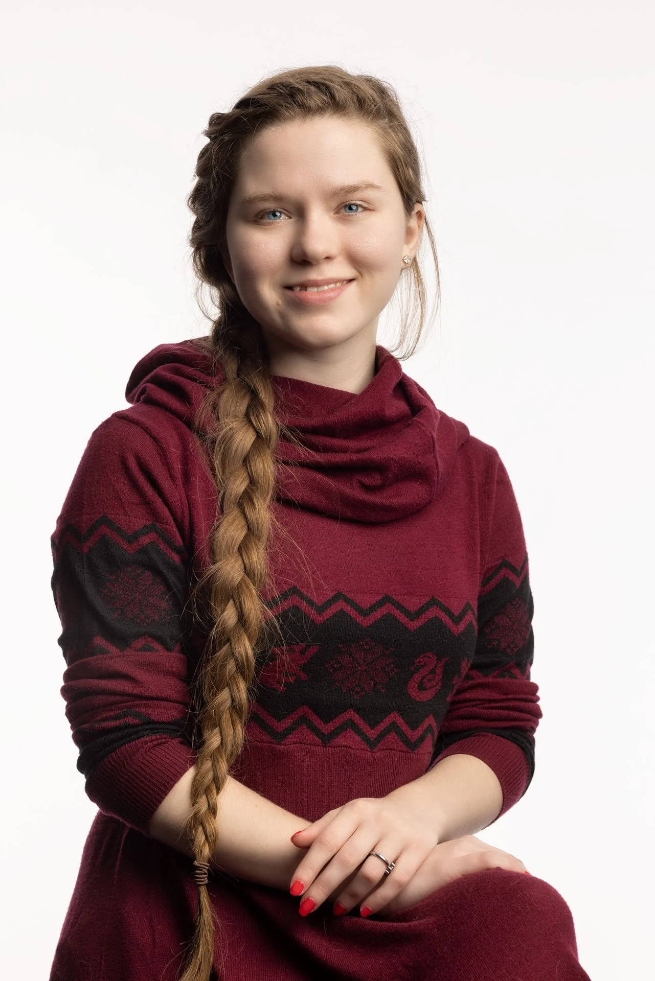 En ung kvinde med langt, lyst hår i en fletning, iført en vinrød sweater med sort mønster, smilende mod kameraet mod en hvid baggrund.