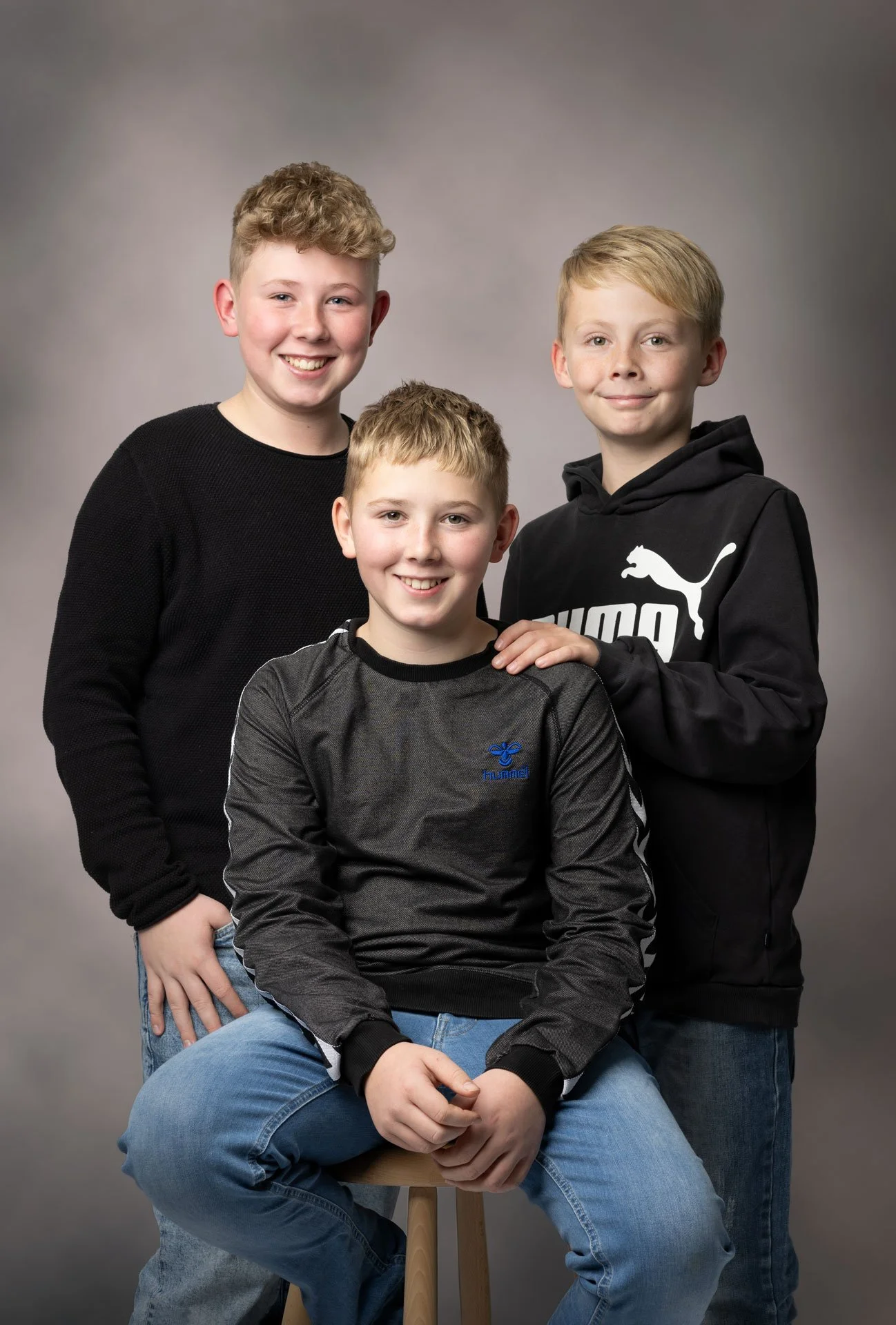 Tre drenge, alle smilende, poseret mod en neutral grå baggrund. En sidder på en skammel, de andre står omkring ham.