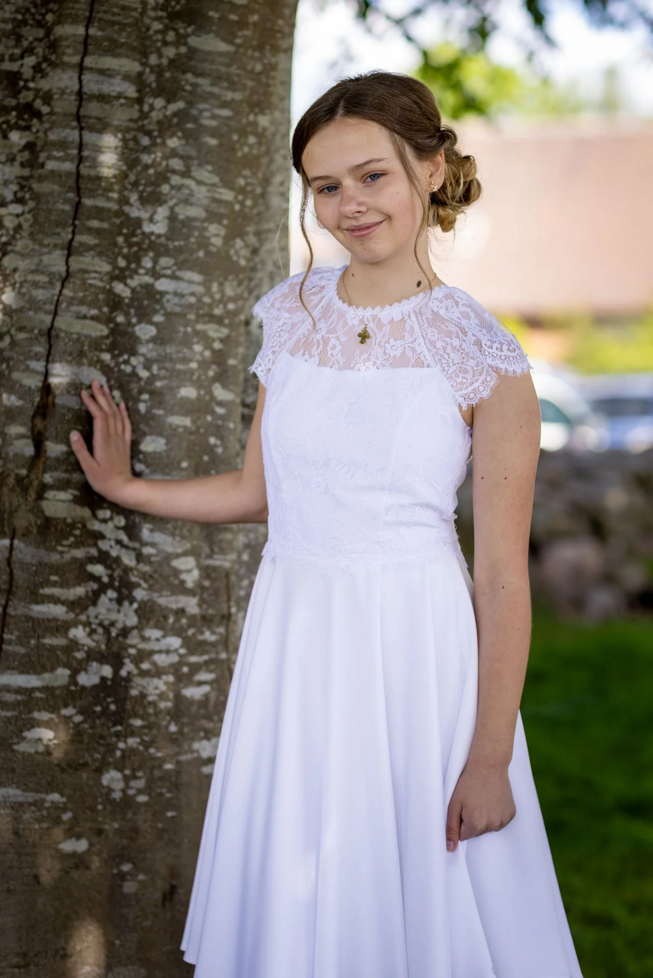 En ung pige i en hvid brudekjole står udenfor ved et træ med grønne blade og smiler.