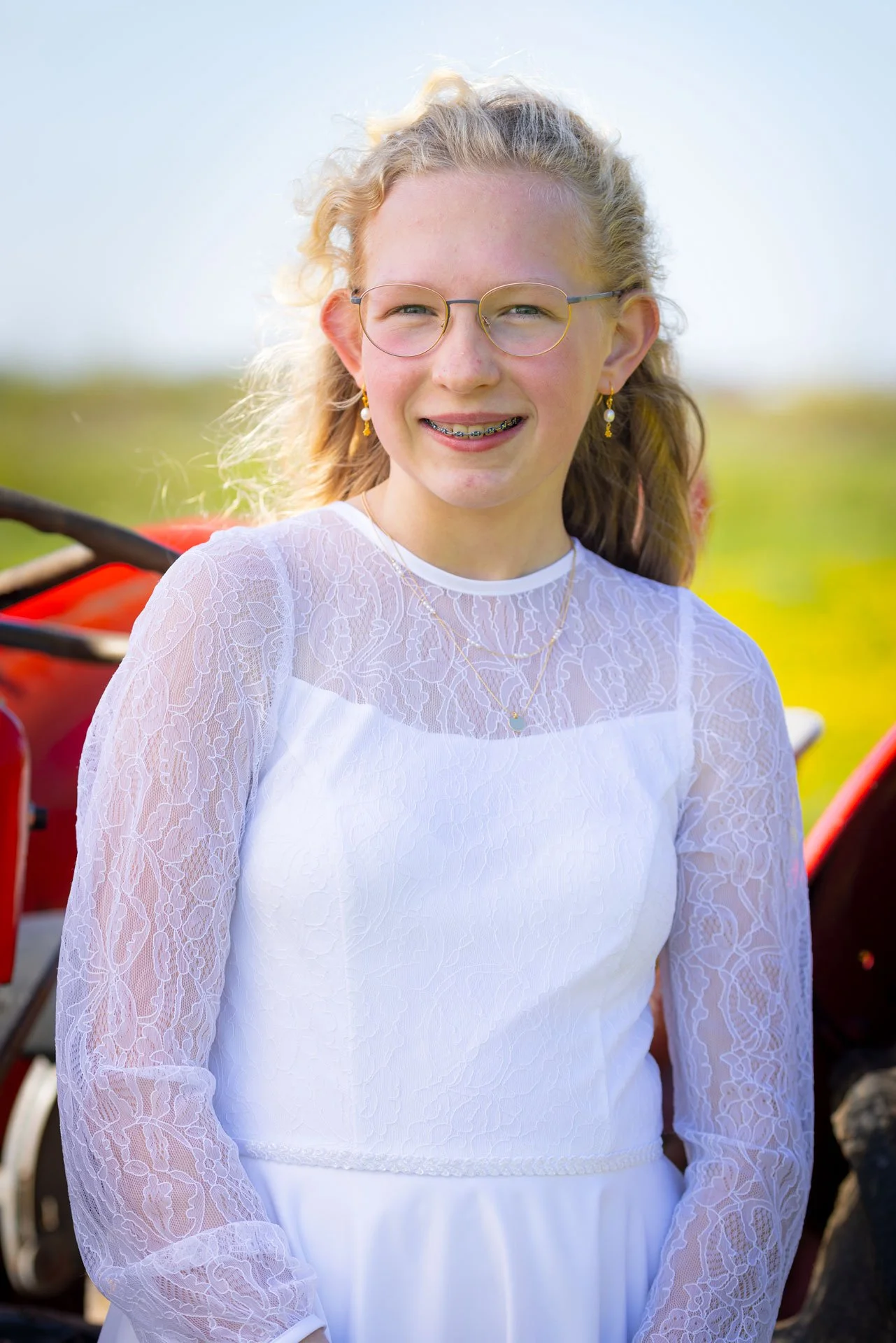 Yngre kvinde med blond hår, briller og smykker, iført hvid brudekjole i udendørs omgivelser.