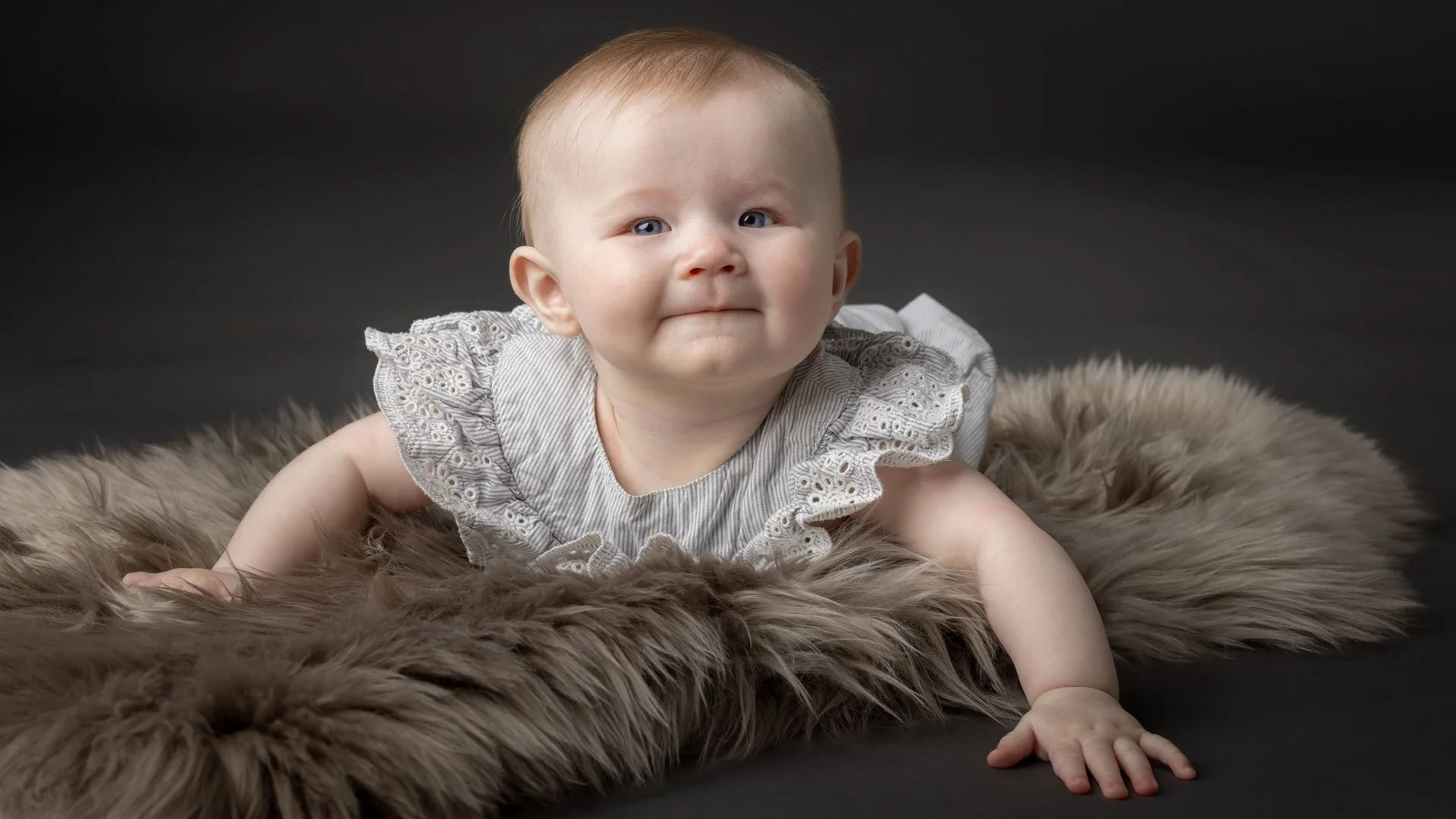 Babyfotografering af en lille pige i lys kjole, der ligger på et blødt pelsunderlag foran en mørk baggrund.