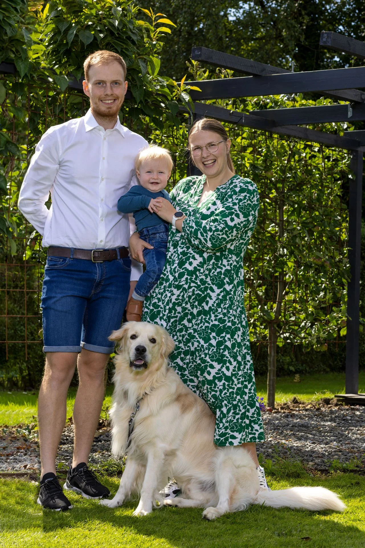 Et familieportræt med tre personer og en hund, fotograferet udendørs med grøn baggrund.