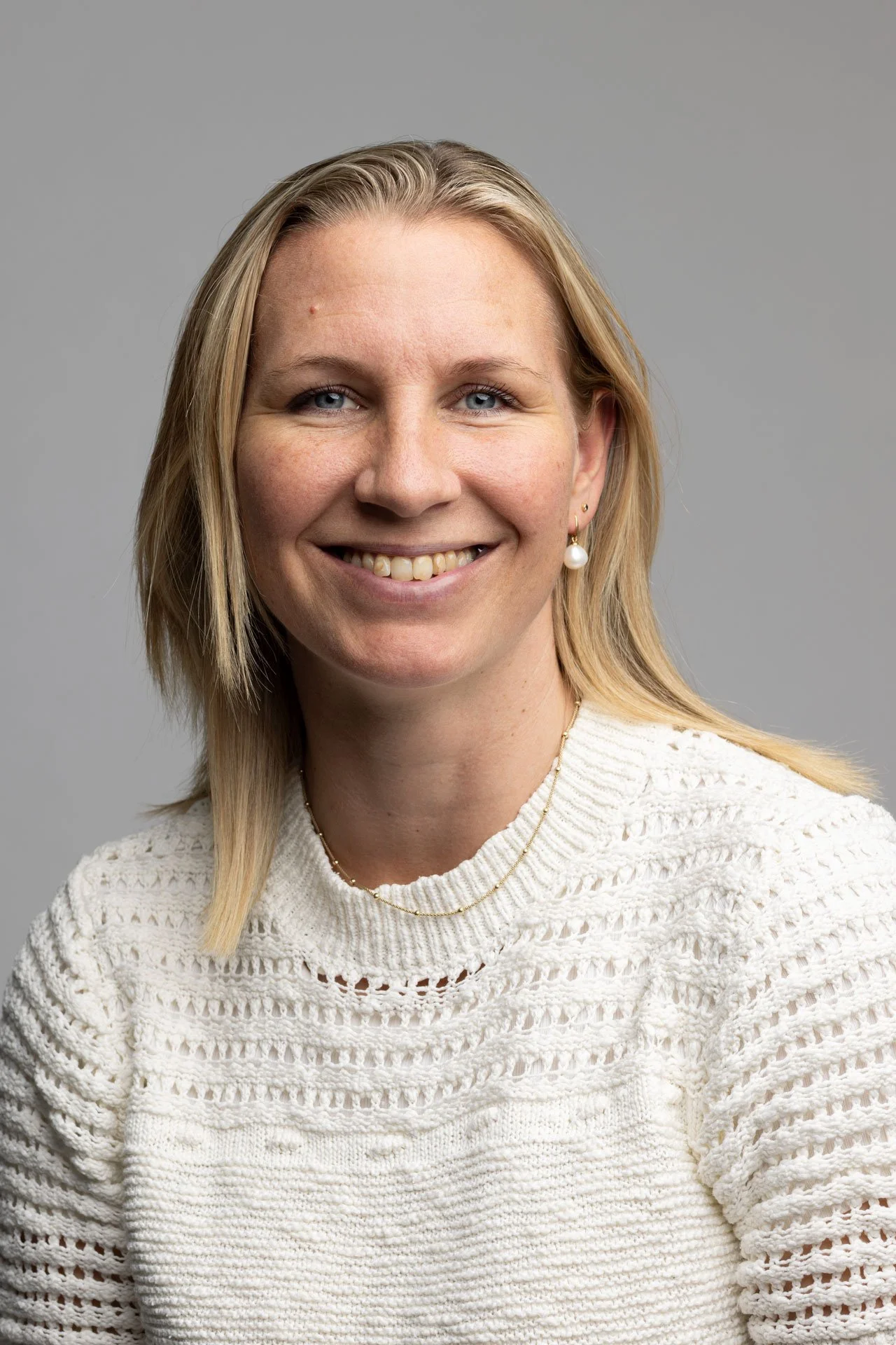 Et portrætbillede af en smilende kvinde med blondt hår, iført en hvid strikket sweater og smykker, mod en neutral grå baggrund.
