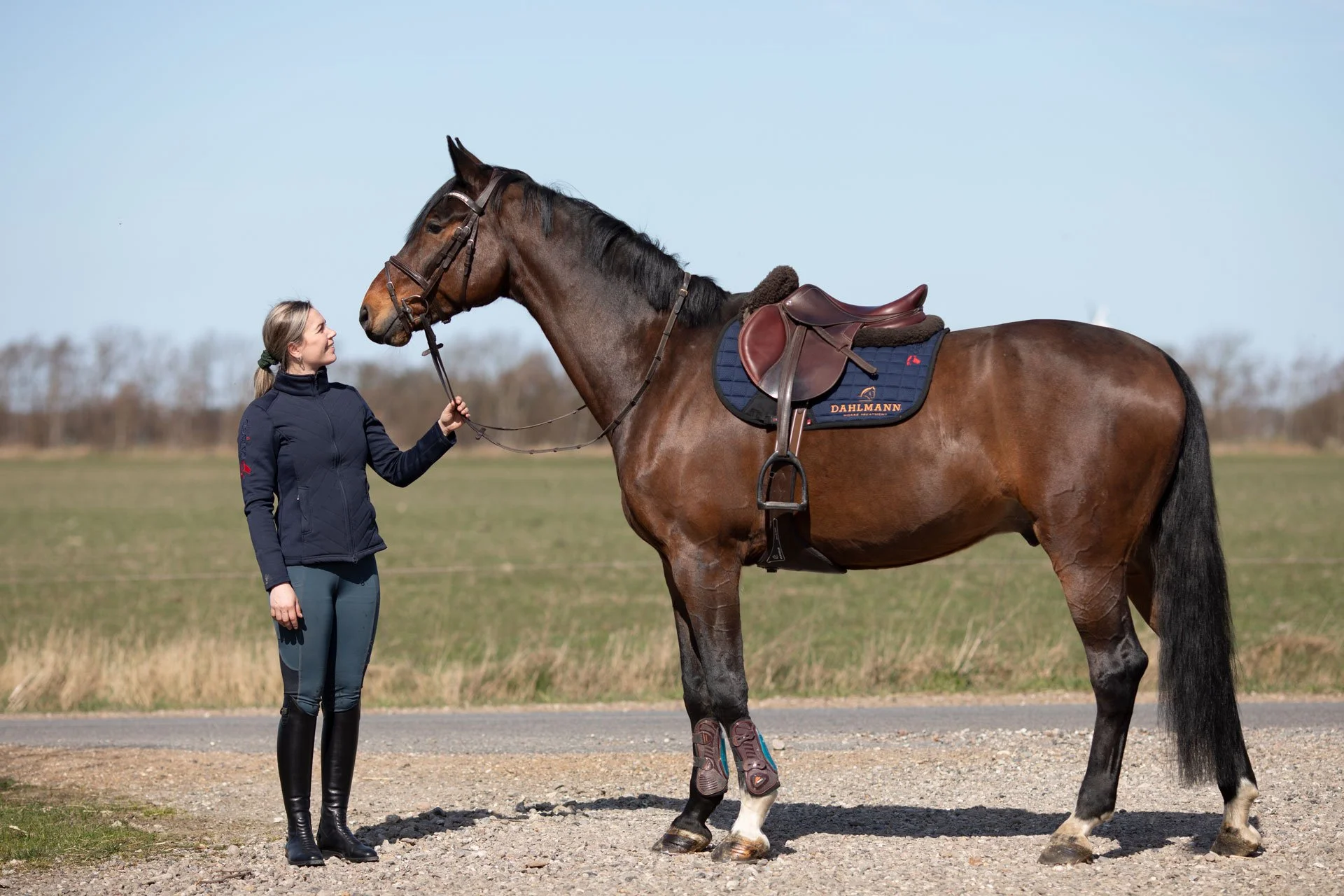En kvinde står foran en brun hest med sadel, på en græsmark på en klar dag.