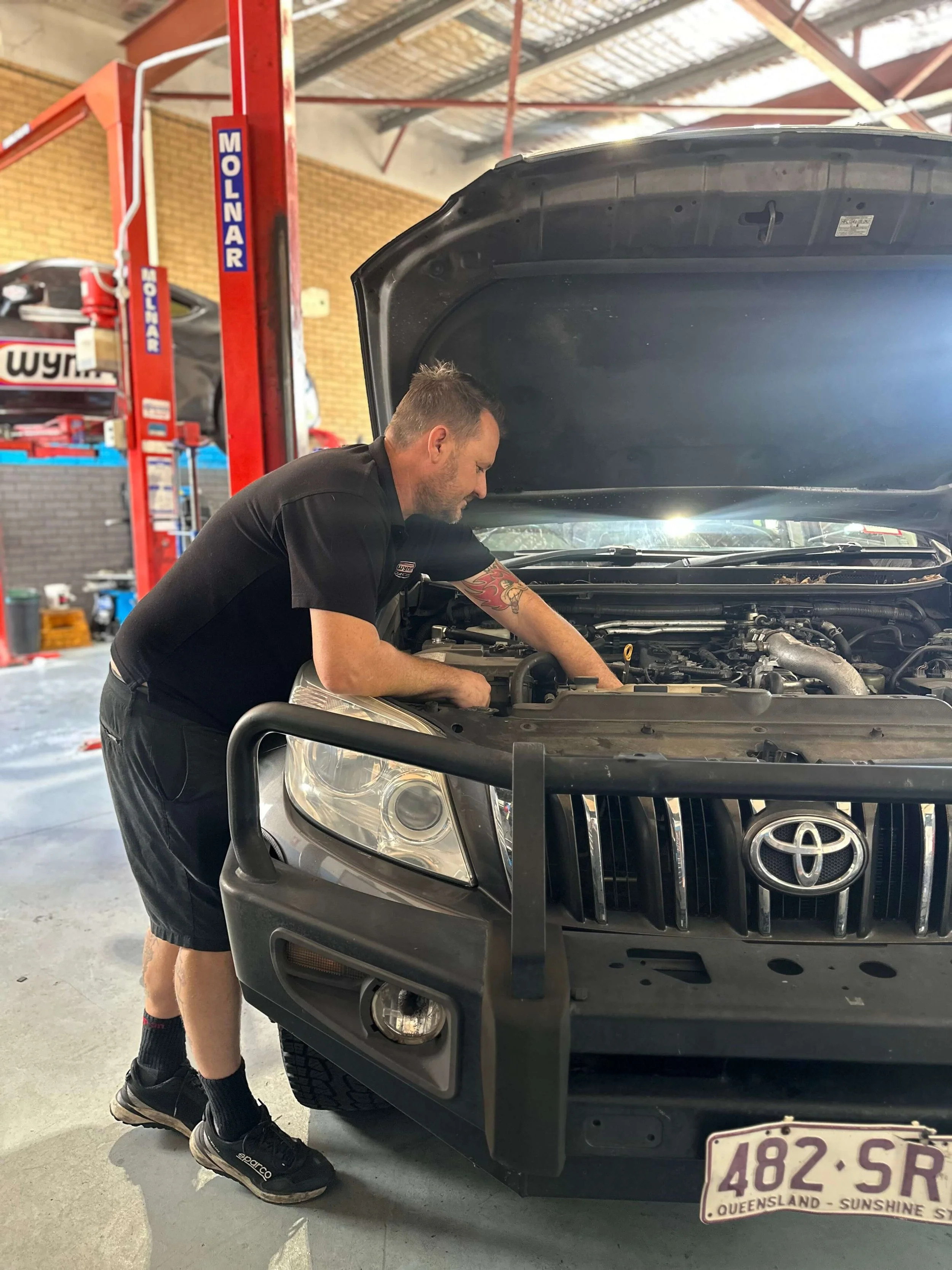 A man working under the hood of a black Toyota vehicle inside a garage or workshop. Car service near me Stafford