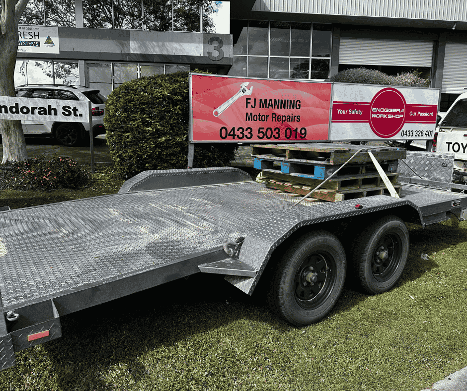 A flatbed trailer parked outdoors displays an advertisement sign for FJ Manning Motor Repairs, with a customer service phone number. The trailer is metal with a diamond plate surface. Trailer roadworthy certificate, Brisbane.