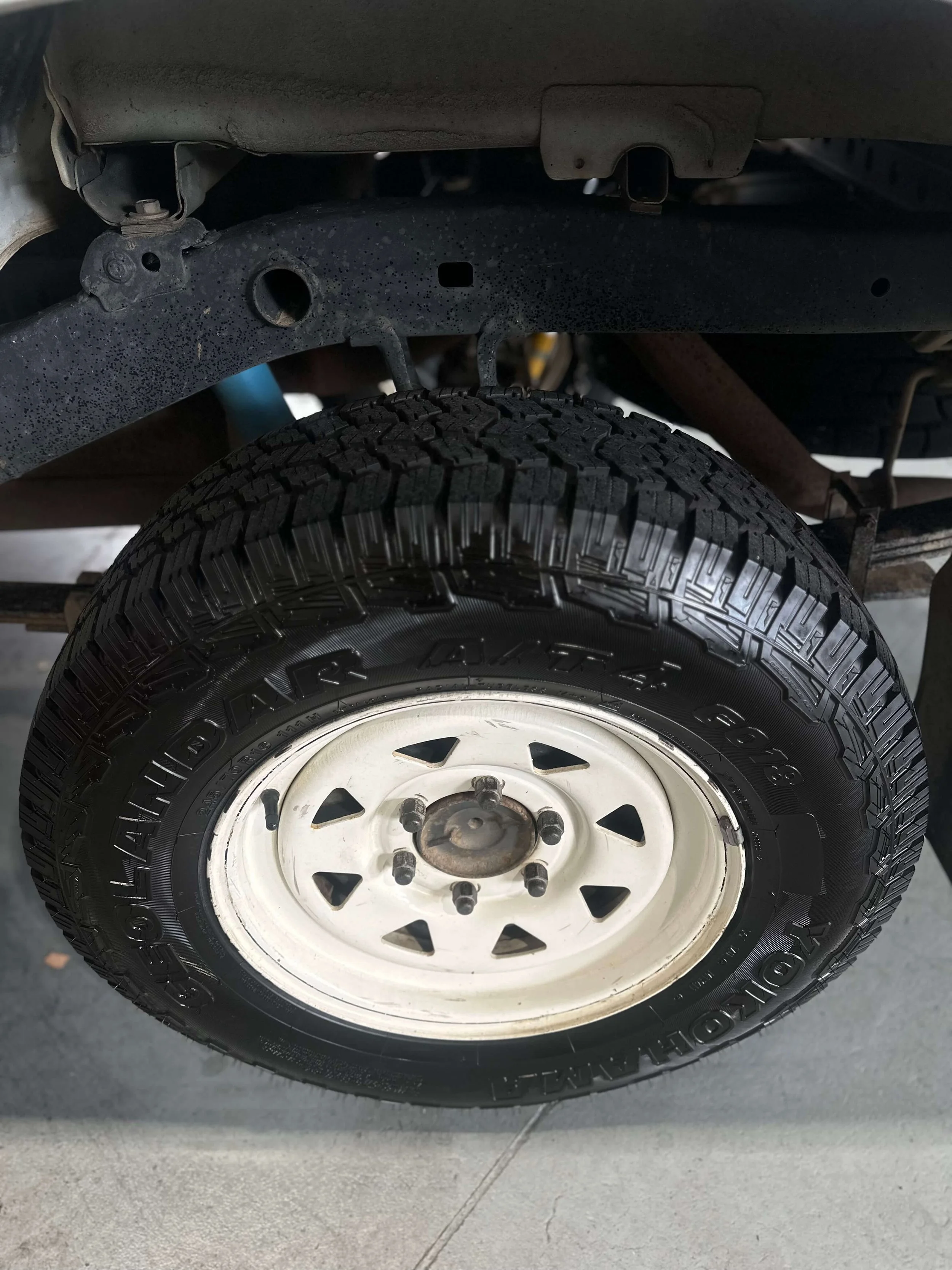 Close-up of a vehicle's tire and wheel with some rust on the rim, under a vehicle's chassis, on a concrete floor. Wheel alignment near me. Tyre repalcement near me.