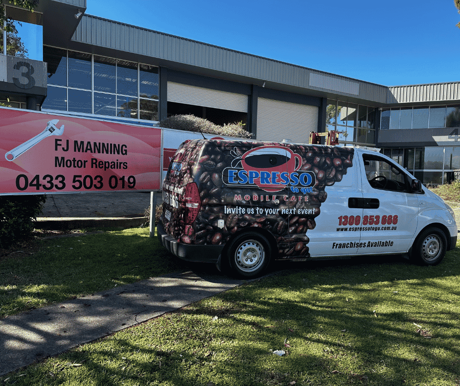 A mobile cafe van with coffee beans design parked on grass, advertising Espresso Go, with a business sign for FJ Manning Motor Repairs nearby, and a modern commercial building in the background. Fleet services Stafford.