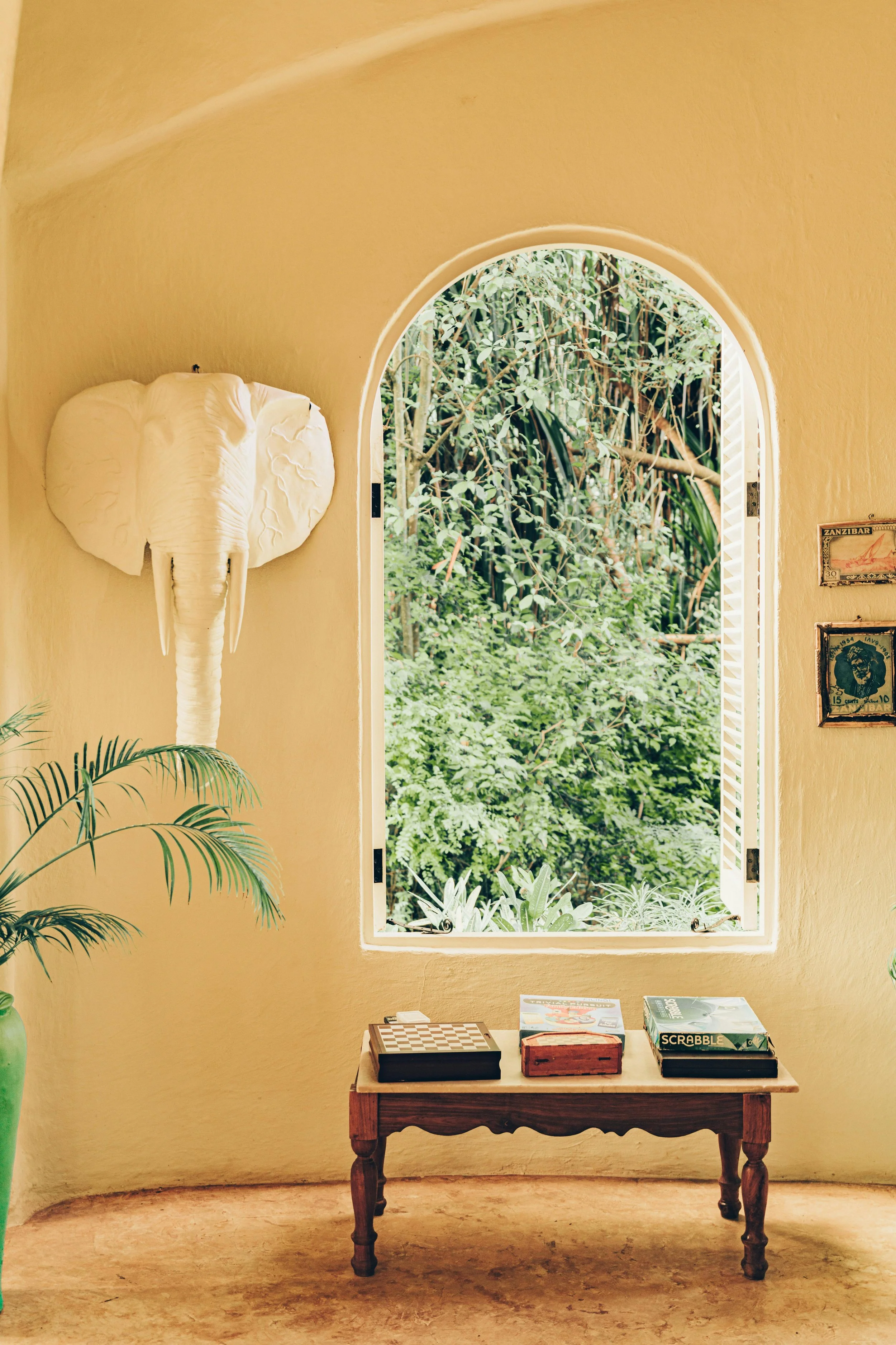 Une pièce avec un mur jaune, une fenêtre en arche ouverte sur une végétation luxuriante, une tête de éléphant en décoration murale blanche, une plante verte à gauche, une petite table en bois avec des jeux de société sur le sol en terre cuite.