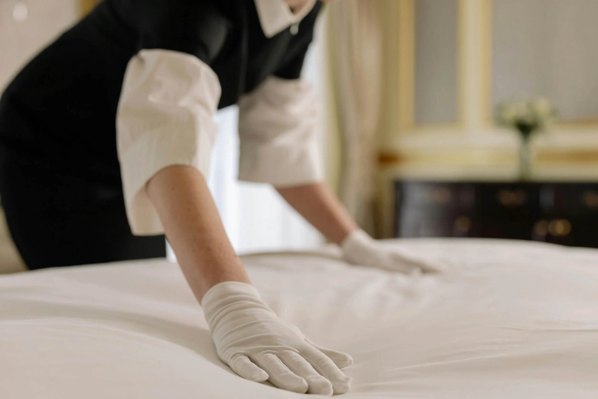 Une personne en tenue de ménage, portant des gants, fait le lit dans une chambre élégante avec un meuble en bois et une fenêtre avec des rideaux