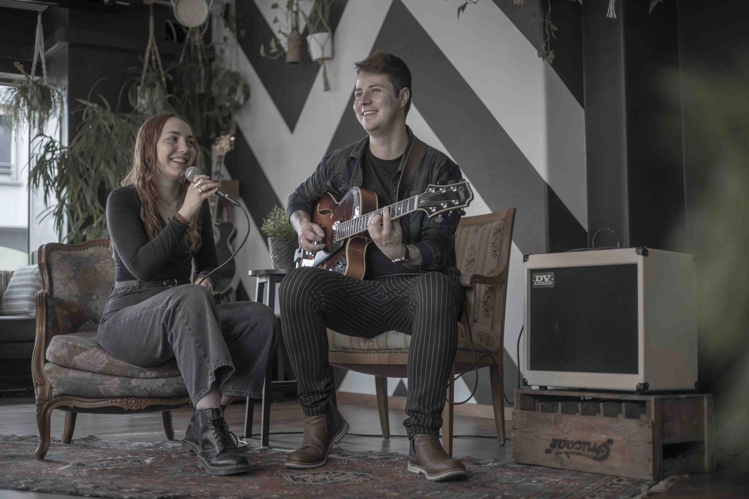 A young woman holding a microphone and a young man playing an electric guitar sit on a vintage-style sofa and a chair in a cozy, modern living room with plants and geometric wall art.