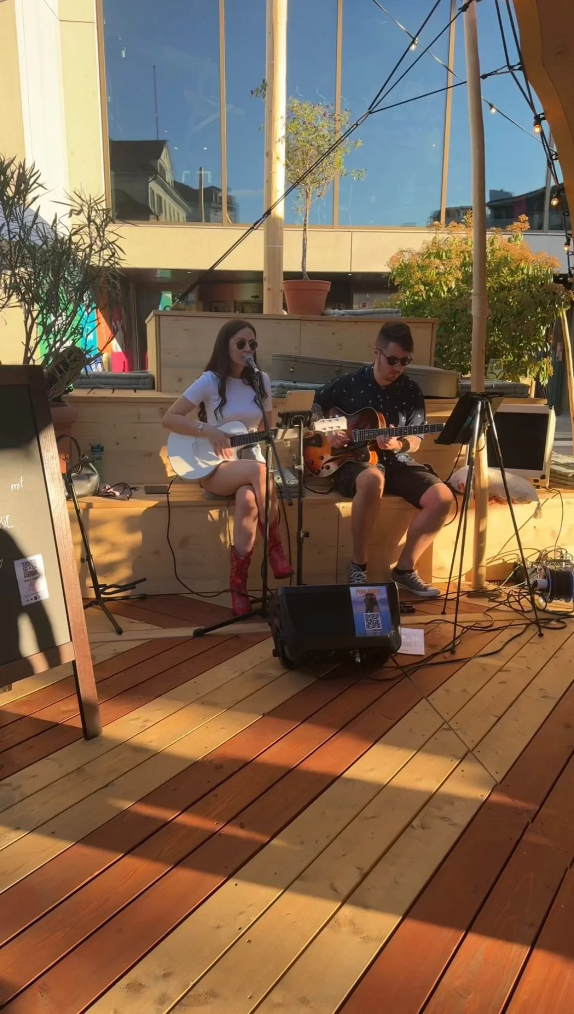 Two musicians performing on a wooden deck outdoor, one woman and one man, both playing acoustic guitars. The woman is singing into a microphone, wearing sunglasses, a white t-shirt, and red boots. The man is wearing sunglasses, a dark shirt, and shorts. There are plants, outdoor furniture, and string lights nearby.