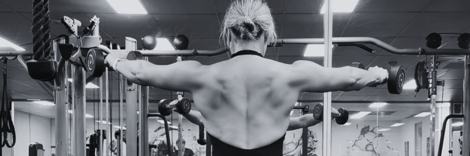 A woman with blonde hair tied back, seen from behind, lifting dumbbells in a gym, with fitness equipment and mirrors in the background.