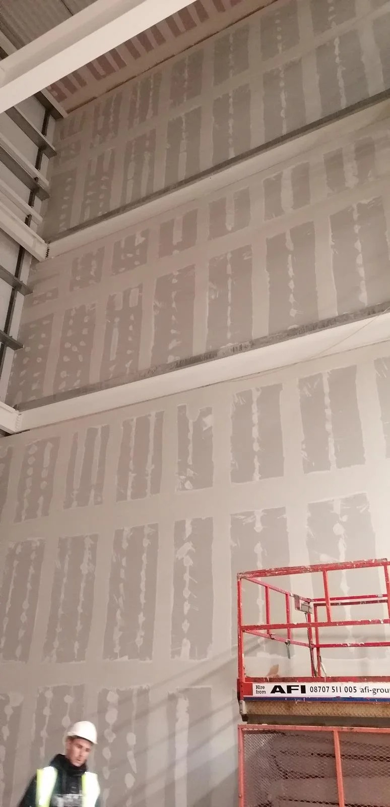 Construction worker wearing a white helmet and reflective vest standing on a red scaffolding in an unfinished building with drywall walls showing joint compound, and an open space to the left with metallic framing.