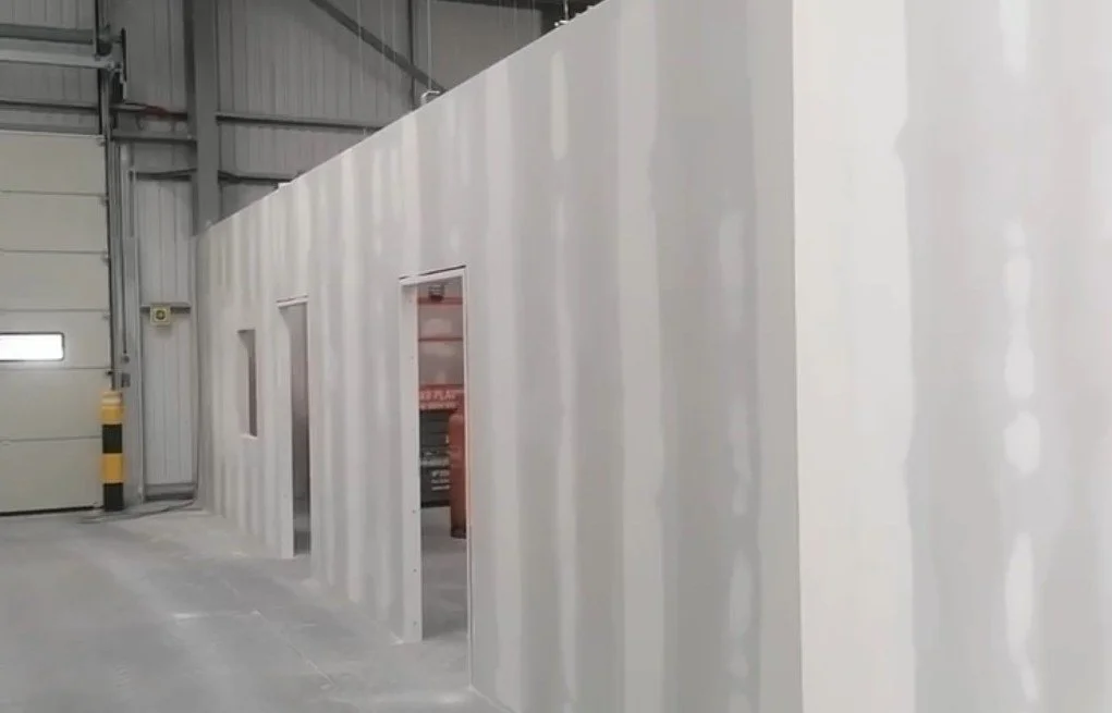 Interior of a large industrial warehouse with a white partition wall featuring three doorways and two small windows, and a garage-style door on the left.