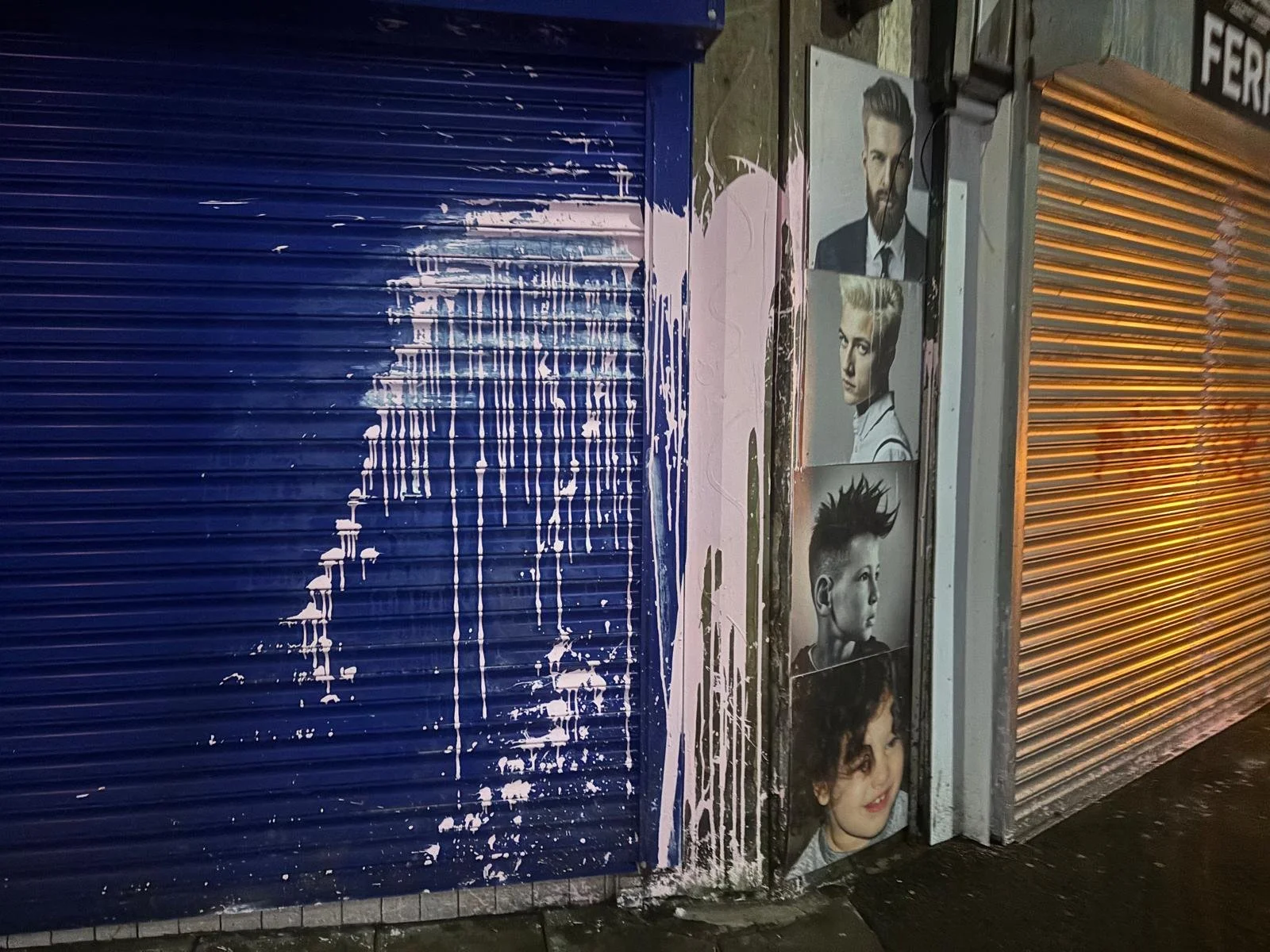 Street scene featuring closed roll-up shutters painted blue and orange, with black-and-white portraits of various hairstyles on a wall adjacent to the shutters.