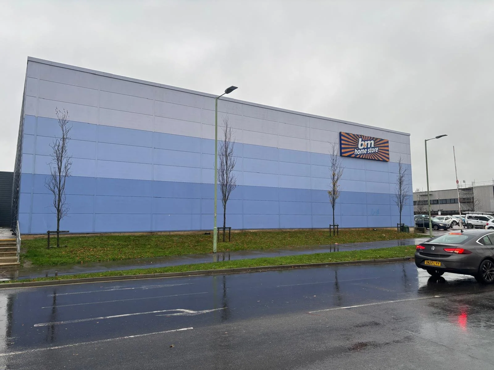 Exterior of a blue building with a sign that reads 'b&m home store' in orange and blue, parked cars, wet pavement, and leafless trees with an overcast sky.
