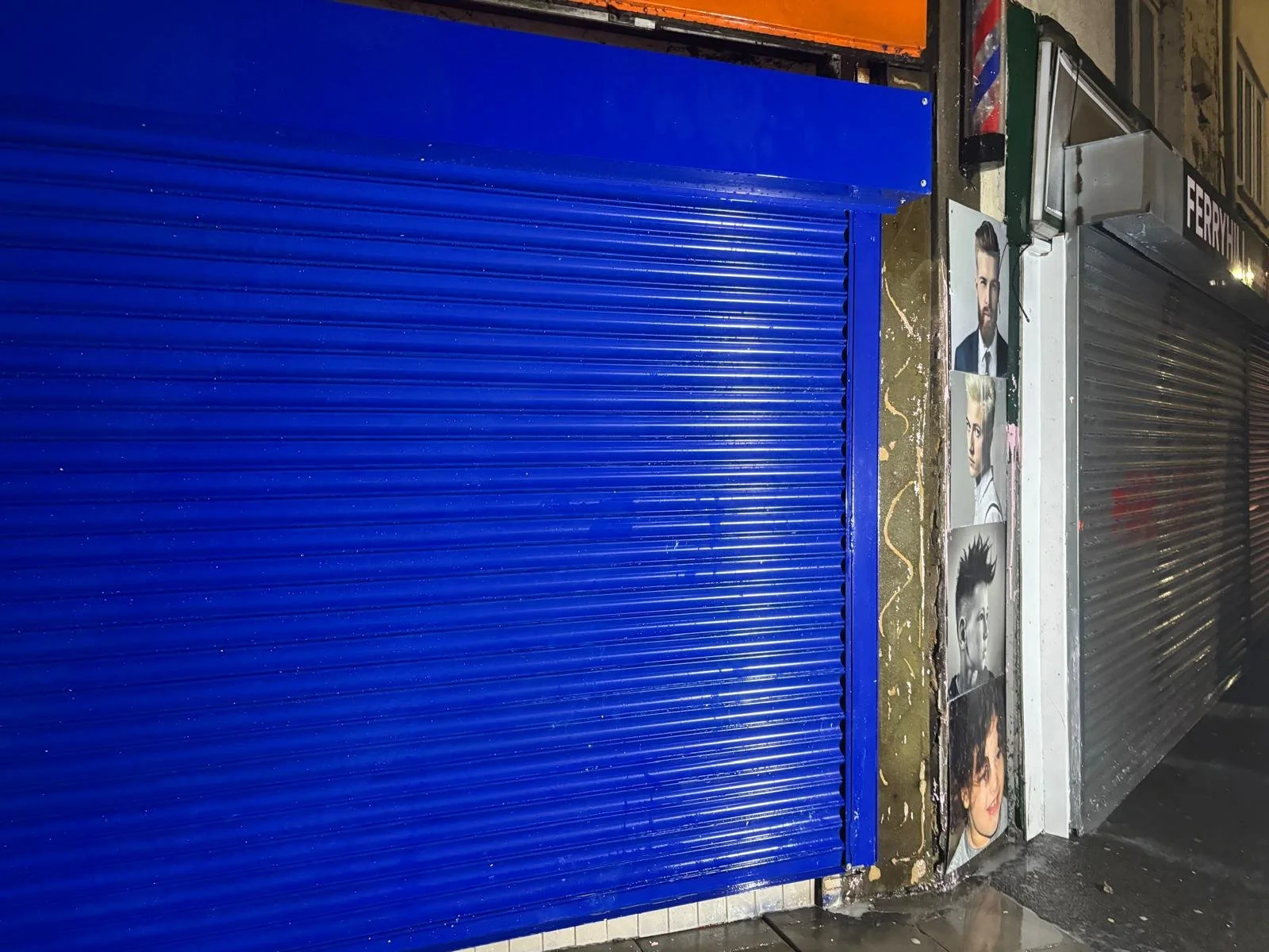 A closed blue metal shutter next to a metallic grey roller shutter. On the sidewalk, there are photos of various hairstyles, including a man with a beard, a woman with short hair, and another woman with longer hair. The shop appears to be a hair salon or barber shop.