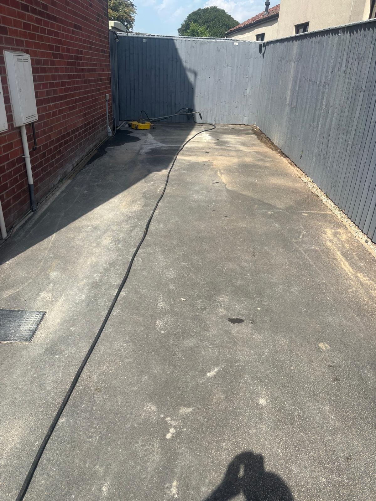 A clean concrete driveway with a power washer and hose, bordered by a brick wall on the left and a wooden fence on the right, with sunlight casting shadows.