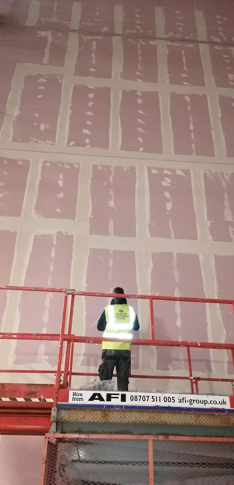 A worker on a red scissor lift painting large pink and white tiles on a wall.