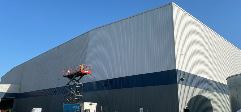 Large industrial building with metal siding and a horizontal blue stripe, featuring a red scissor lift parked outside under a clear blue sky.