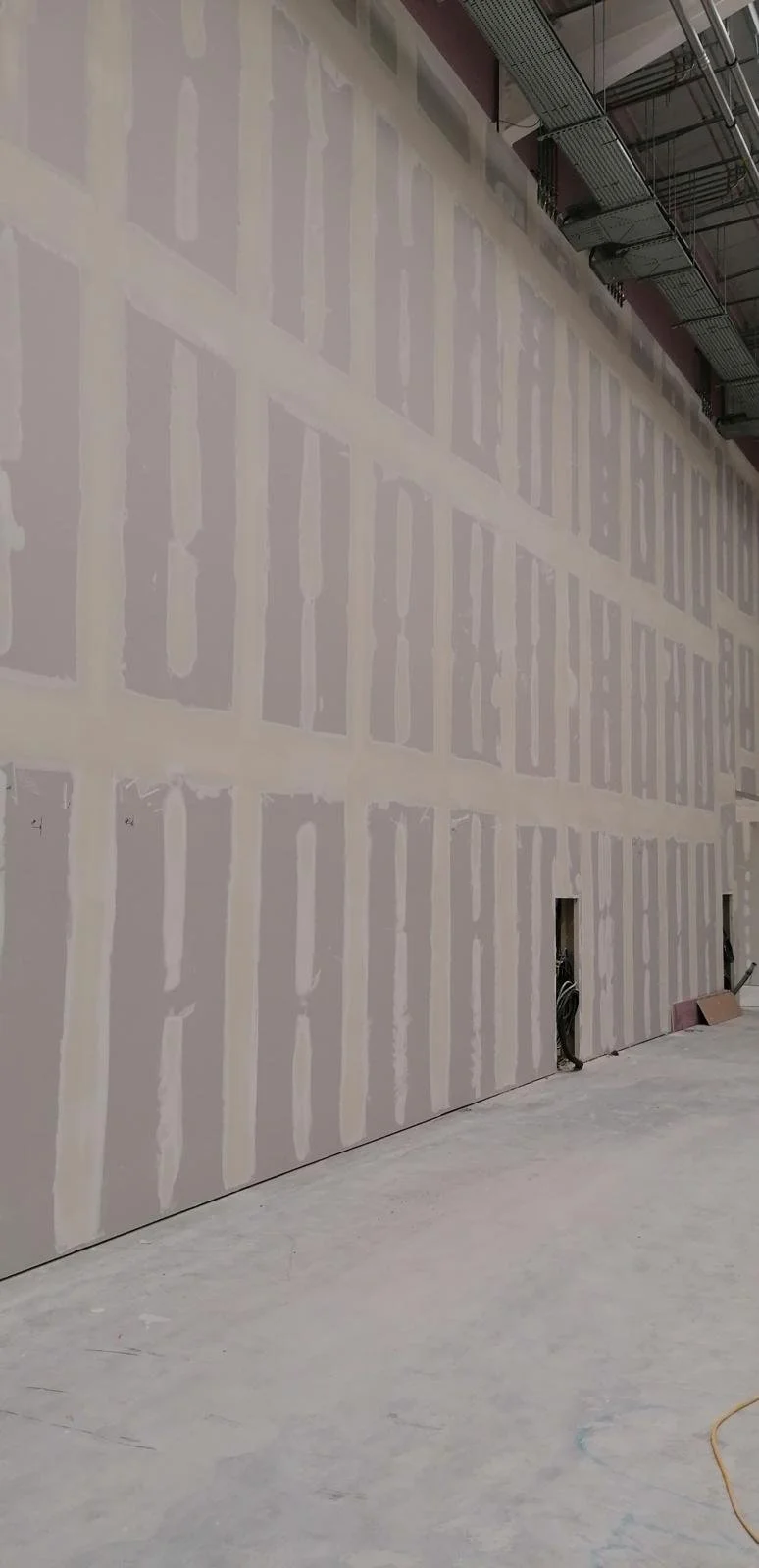Interior of a building under construction with a large drywall wall showing taped and mudded seams. The floor is unfinished concrete, and construction materials and wires are visible.