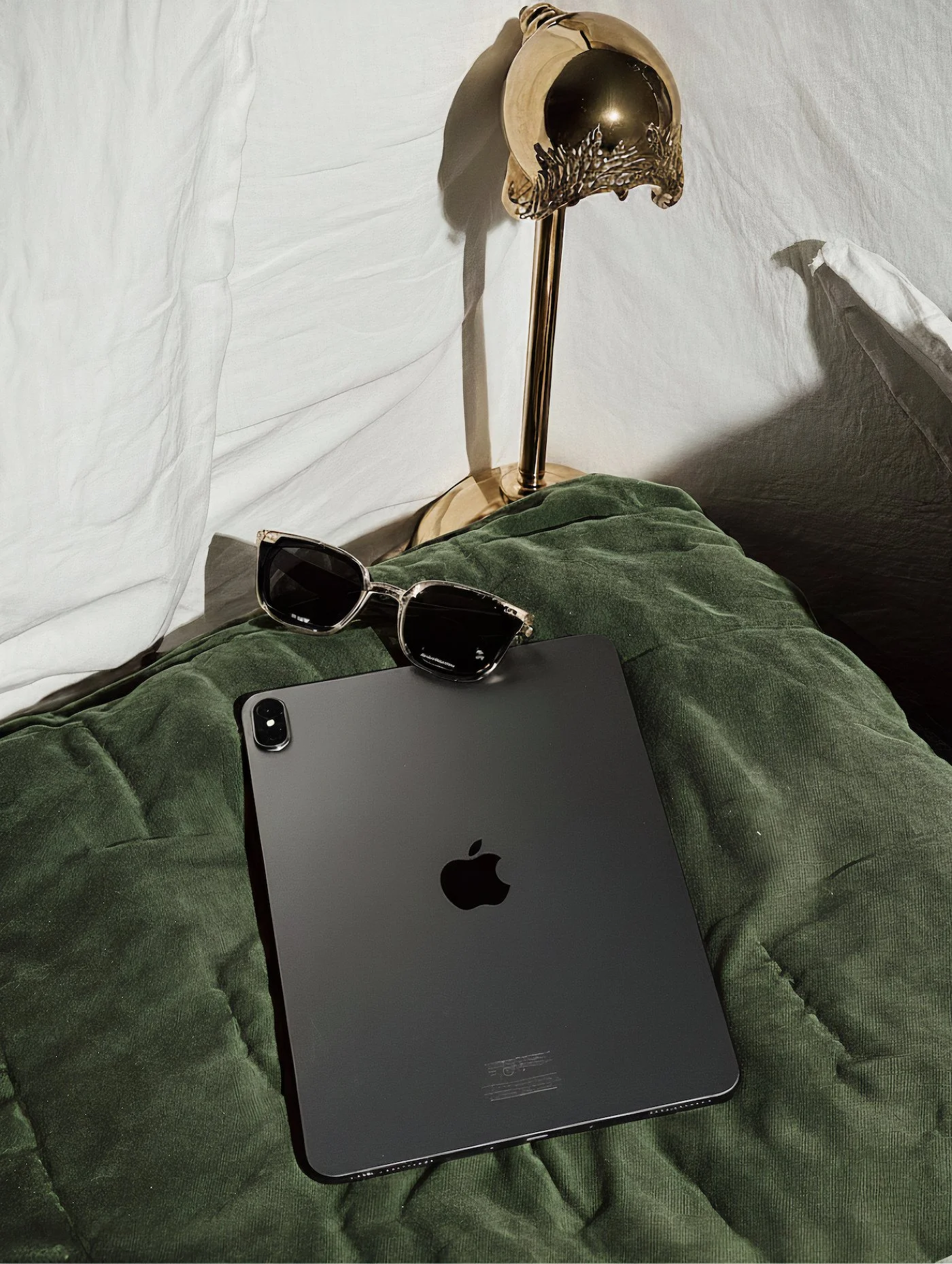 Black iPad with Apple logo, sunglasses, and a gold-colored lamp with a decorative shade on a green velvet surface with white bedding in the background.