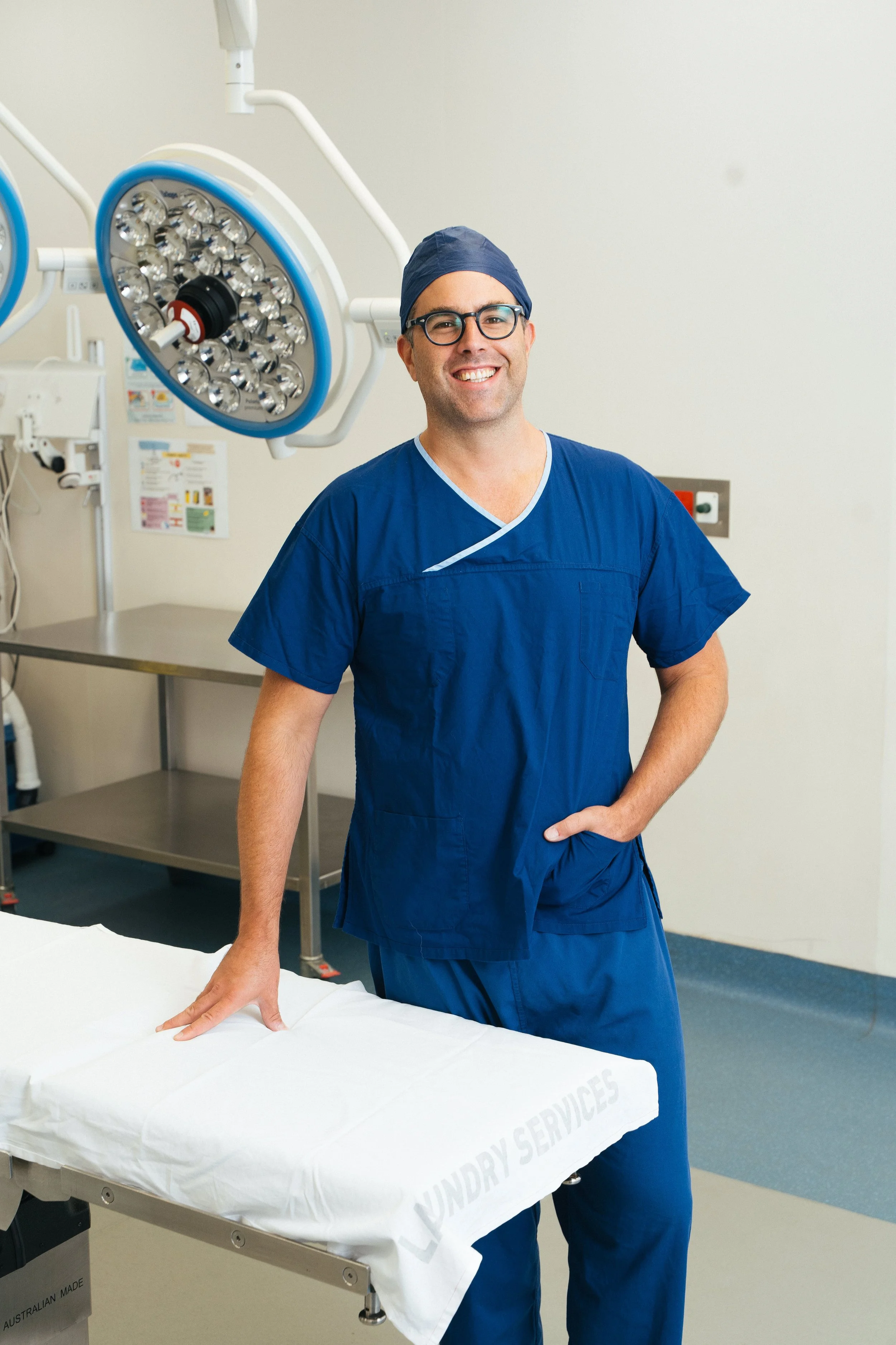 A smiling male healthcare professional in blue scrubs and a cap standing in a hospital or operating room.