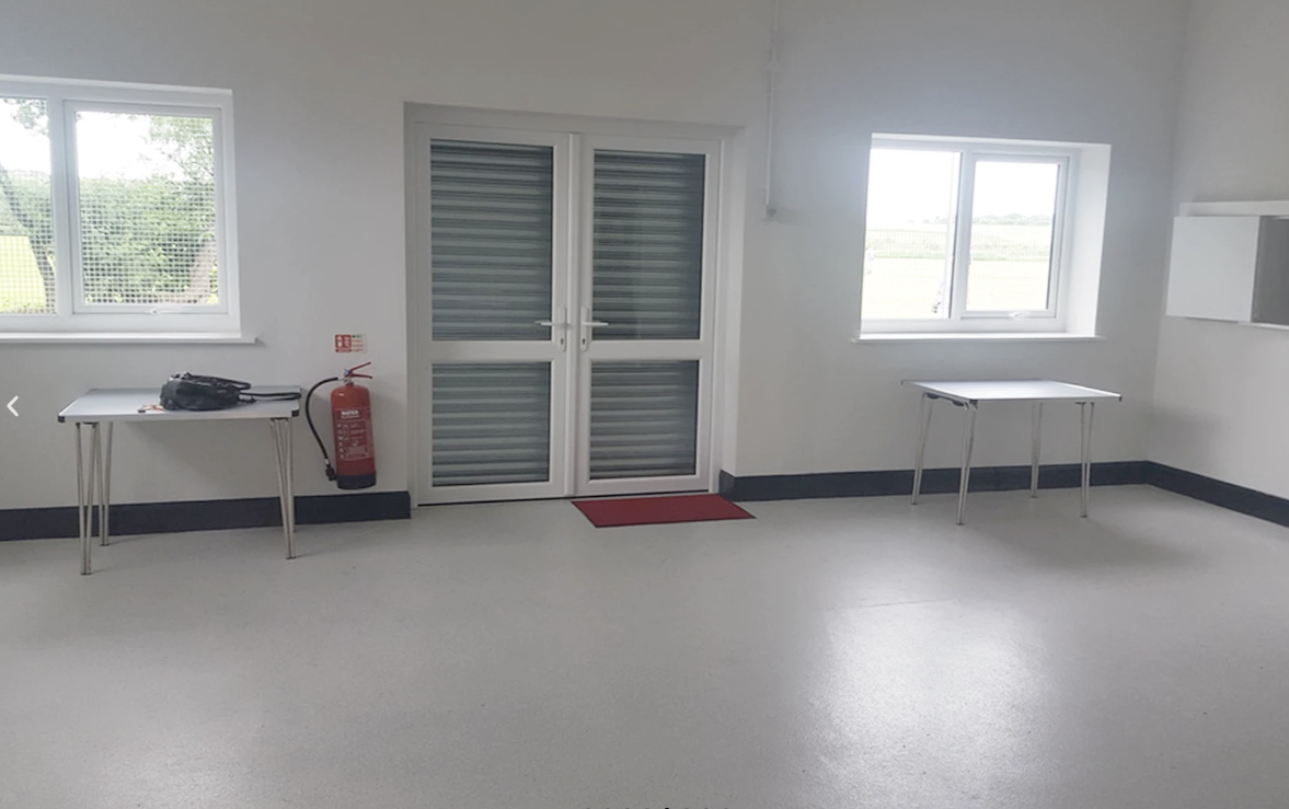 Empty room with white walls, gray floor, two windows, a door with a red mat, and two tables, one on each side, with a bag and a fire extinguisher nearby.