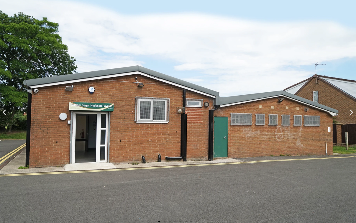 Brick building with a sign that reads 'William Segar Hodgson Pavilion' and a green door, windows with metal grates, situated beside a paved road with yellow line markings.