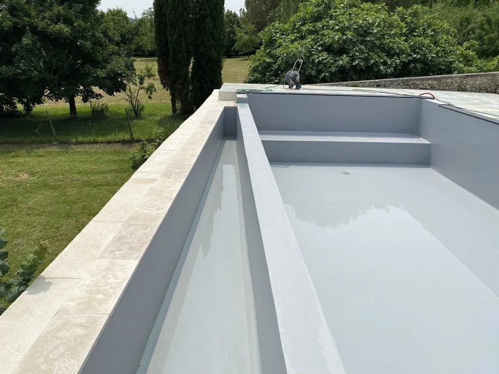 Piscine vide avec margelle en pierre, située dans un jardin entouré d'arbres et de buissons, sous un ciel clair.